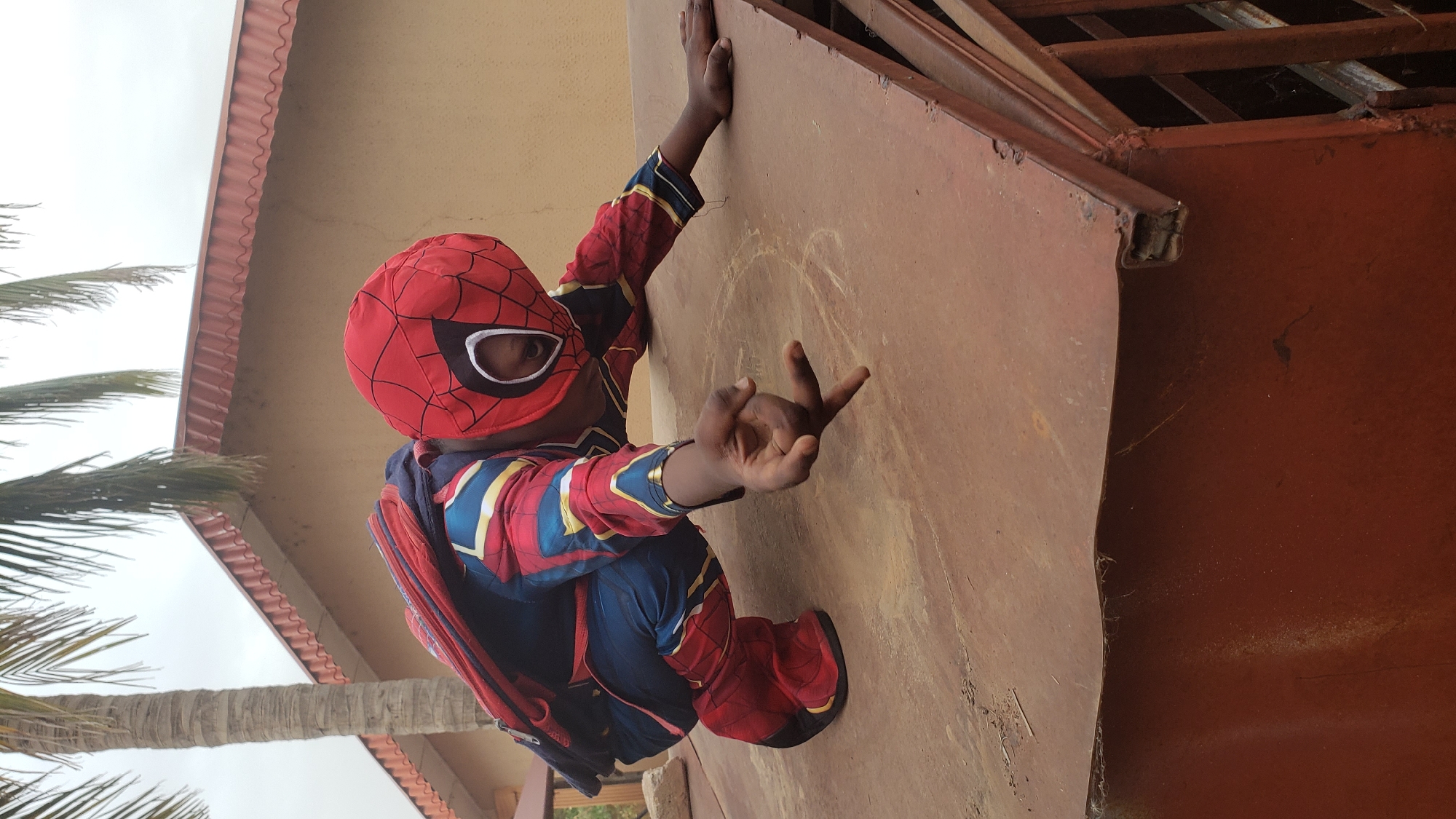 A child in a Spider-Man costume is positioned as if climbing a rusty, brown metal structure. The child wears a red and blue Spider-Man suit with a red mask and white eye patches. They are holding onto the metal structure with one hand and making a gesture with the other. A palm tree is visible to the left, suggesting a tropical or semi-tropical environment. A beige building with a red-tiled roof is partially visible in the background. The location is Sabon Tasha, Nigeria.