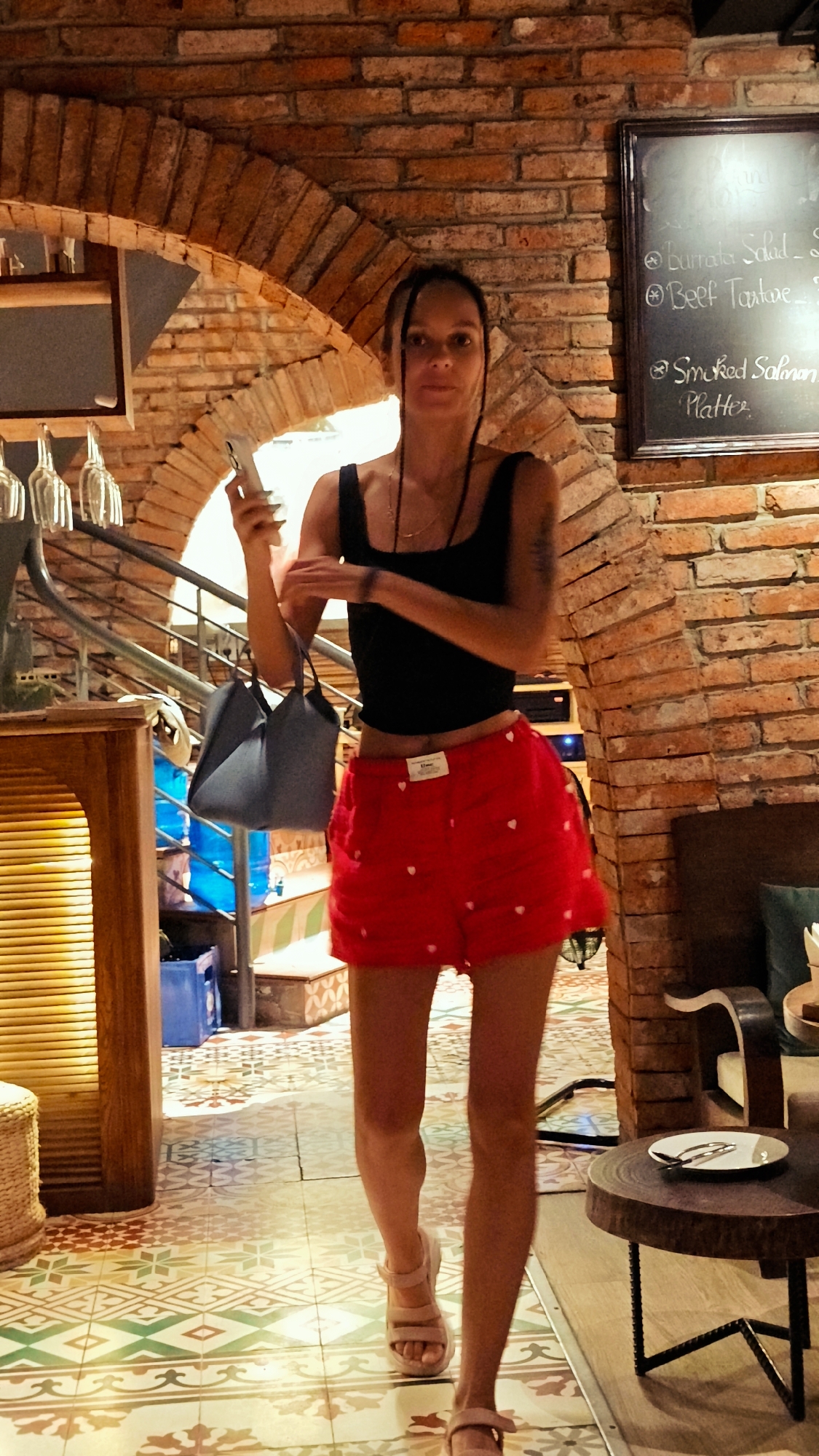 The image depicts a young woman walking towards the camera in a well-lit, indoor establishment with exposed brick walls and arched doorways. She is wearing a black tank top and red shorts with white heart patterns. She is holding a smartphone in her left hand and carrying a grey handbag on her left arm. Her hair is pulled back in braids. The floor is tiled with a colorful, geometric pattern. A blackboard menu is visible on the brick wall to her right, displaying food items in English. In the background, there are stairs, a wooden counter, and glassware hanging from a rack. The overall ambiance suggests a restaurant or bar in Hanoi, Vietnam, given the style of the brickwork, tiling, and the presence of a menu with common Western dishes.