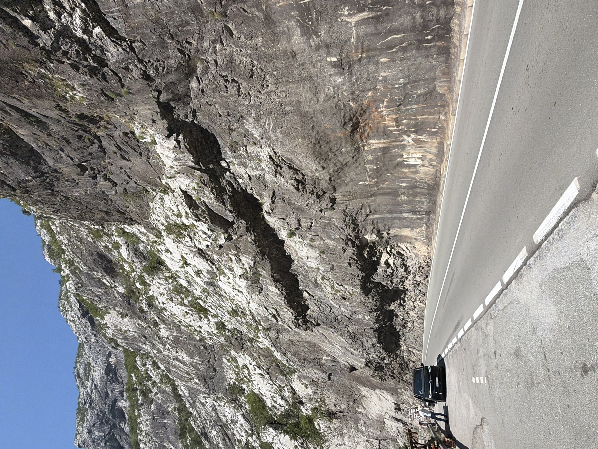 The image captures a section of the M2 highway running alongside a sheer, rugged mountain face on a clear, sunny day. The mountain is characterized by its grey, rocky texture, with patches of white mineral deposits and sparse green vegetation clinging to its steep inclines. The road is paved and well-maintained, with white dashed lines marking the lanes and solid white lines delineating the shoulder.
On the left side of the road, a black van is parked. A person, appearing to be a man, stands beside the van, looking away from the camera. Further left, a small wooden structure resembling a market stall is visible, with what appear to be goods displayed on it. Another person, partially obscured, stands near the stall. The ground on the shoulder is a mixture of asphalt and gravel.
The mountain face itself shows signs of erosion and weathering, with streaks of rust-colored discoloration and some graffiti-like markings etched into the rock in the lower right portion of the image. The sky is a clear, vibrant blue, indicating fair weather. The scene appears to be on the outskirts of Vlahovići, Montenegro, a region known for its dramatic mountain landscapes and scenic roadways.