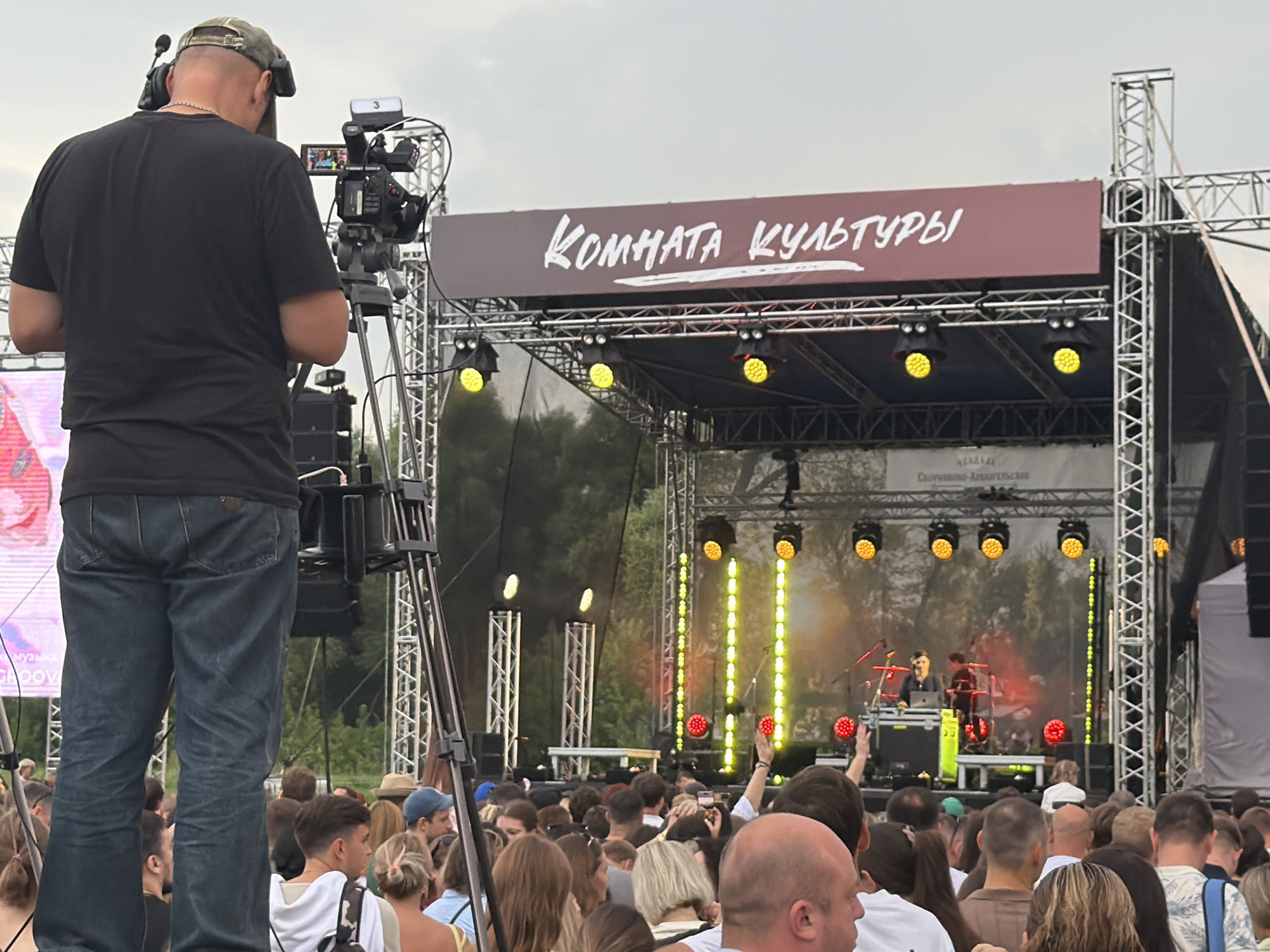 The image shows an outdoor music festival. In the foreground, a cameraman is filming the stage with a professional video camera. He is wearing a black t-shirt and jeans. Behind him, a large crowd of people are watching the performance. The stage has a banner that reads "КОМНАТА КУЛЬТУРЫ" (Room of Culture). There are several spotlights and stage lights on the stage, with a band performing. The event appears to be taking place in the city of Скорняково, Russia, as indicated by the presence of signage and the overall atmosphere of a local gathering. The sky is overcast, suggesting potentially changeable weather.