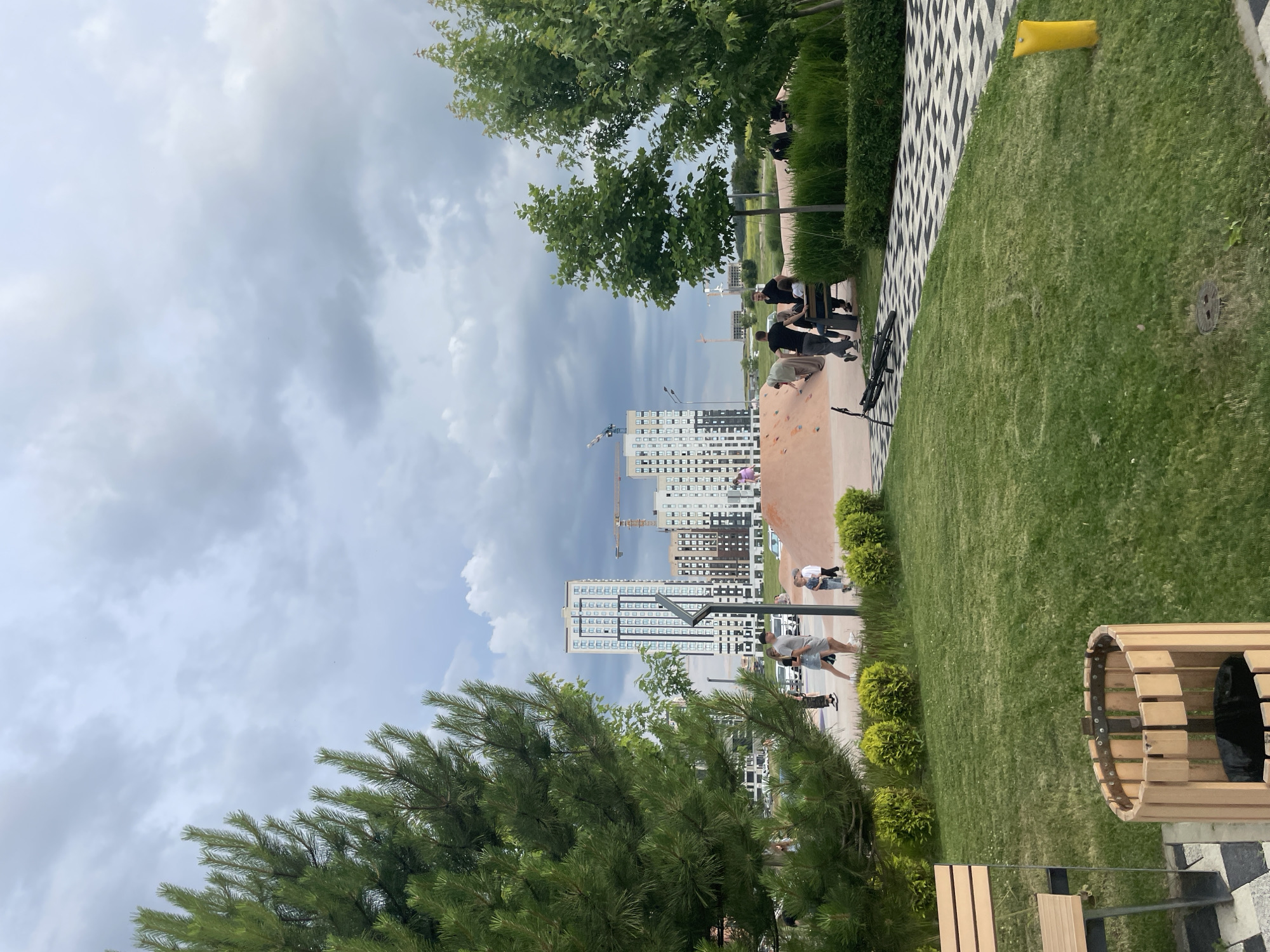 The image depicts a public park area with a playground featuring a sand dune structure. Several individuals, including adults and children, are present in the park. Some are walking, while others appear to be supervising children on the playground. In the background, modern high-rise buildings are visible, suggesting an urban setting. Trees and green landscaping are present throughout the park. A bicycle is lying on the ground near a pathway. The sky is overcast with clouds. This scene is located in the city of Stavropol, Russia, possibly within a newly developed residential area with integrated recreational spaces.