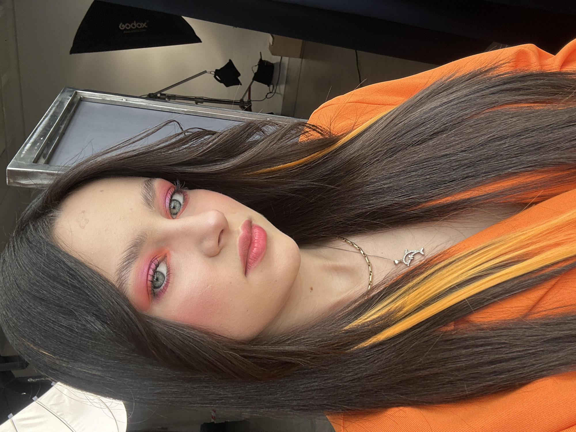 A close-up, angled shot shows a young woman with dark brown hair and dyed yellow streaks. She is wearing a bright orange top and a delicate gold necklace with a dolphin pendant. Her makeup features bright pink eyeshadow and blush, and her lips are a soft pink. The background is cluttered with studio lighting equipment, including a Godox softbox and reflectors, suggesting a professional photoshoot setting. The location appears to be indoors, and there is no evidence to confirm it is in Yerevan, Armenia.