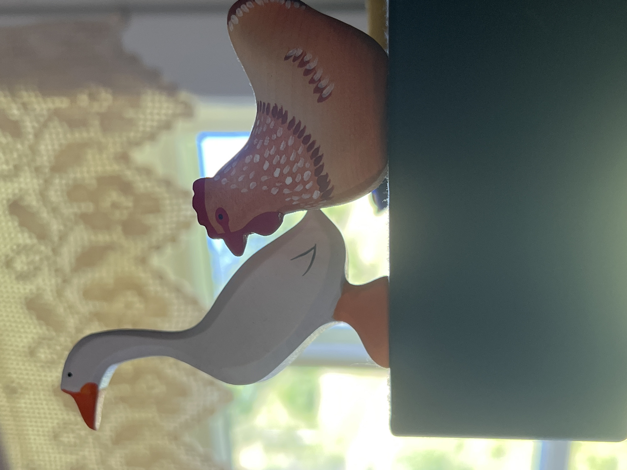 A still-life photograph shows two wooden bird figures, a goose and a hen, positioned on a dark green surface. The goose, to the left, is predominantly white with a gray neck and orange beak and feet. The hen, to the right, is a light brown color with red comb and wattles, and features white and reddish-brown painted feather details. Both figures are positioned in profile, facing towards the right side of the frame. In the background, a window with a lace curtain is visible, allowing natural light to illuminate the scene and cast faint shadows. The overall environment suggests an indoor setting, possibly a windowsill or shelf. There are no people present in the image.