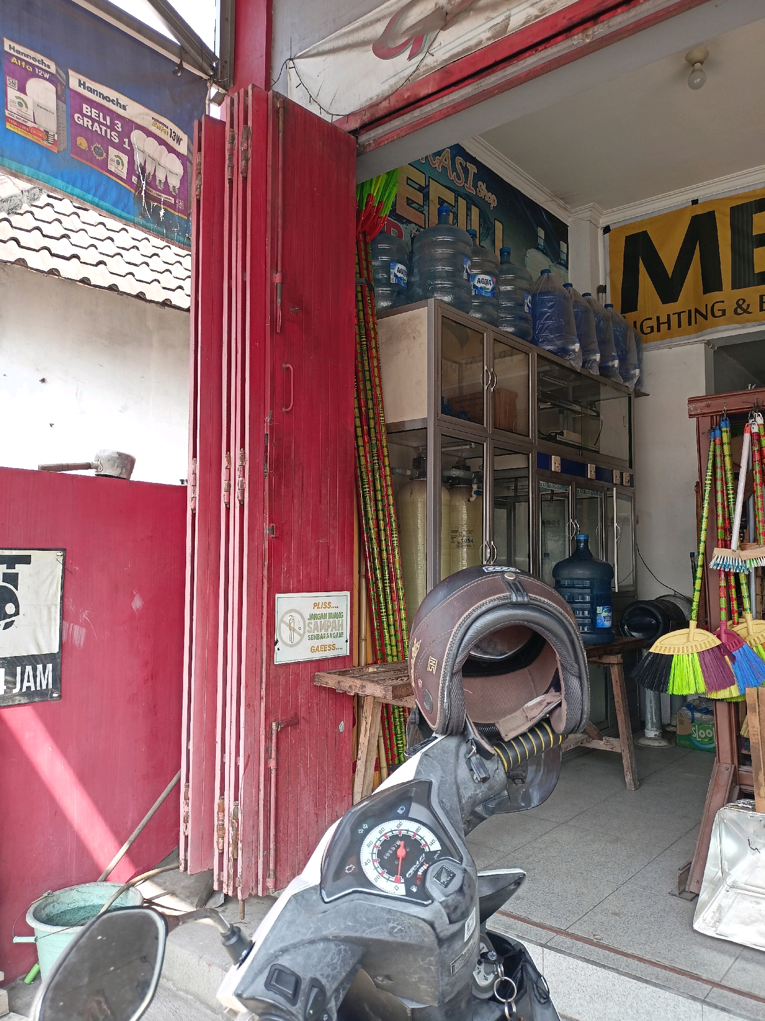 The image depicts the exterior of a shop in Payaman, Indonesia, on a sunny day. The shop appears to be a retail establishment selling bottled water and possibly other household items. In the foreground, a motorcycle is parked, with a brown helmet placed on its handlebars.
The shop's entrance features red, folding metal doors, partially open to reveal the interior. Inside, shelves are stacked with numerous large bottles of drinking water. To the right of the entrance, cleaning supplies such as brooms and dustpans are displayed. A yellow banner with black text, possibly indicating the shop's name or services, is visible above the cleaning supplies. A smaller sign is attached to the red doors, displaying a message in Indonesian with an image of a person discarding trash.
In the background, a white building with a tiled roof is partially visible. The overall scene suggests a commercial area in a local Indonesian town. The lighting indicates it is daytime. No people are directly visible in the image.