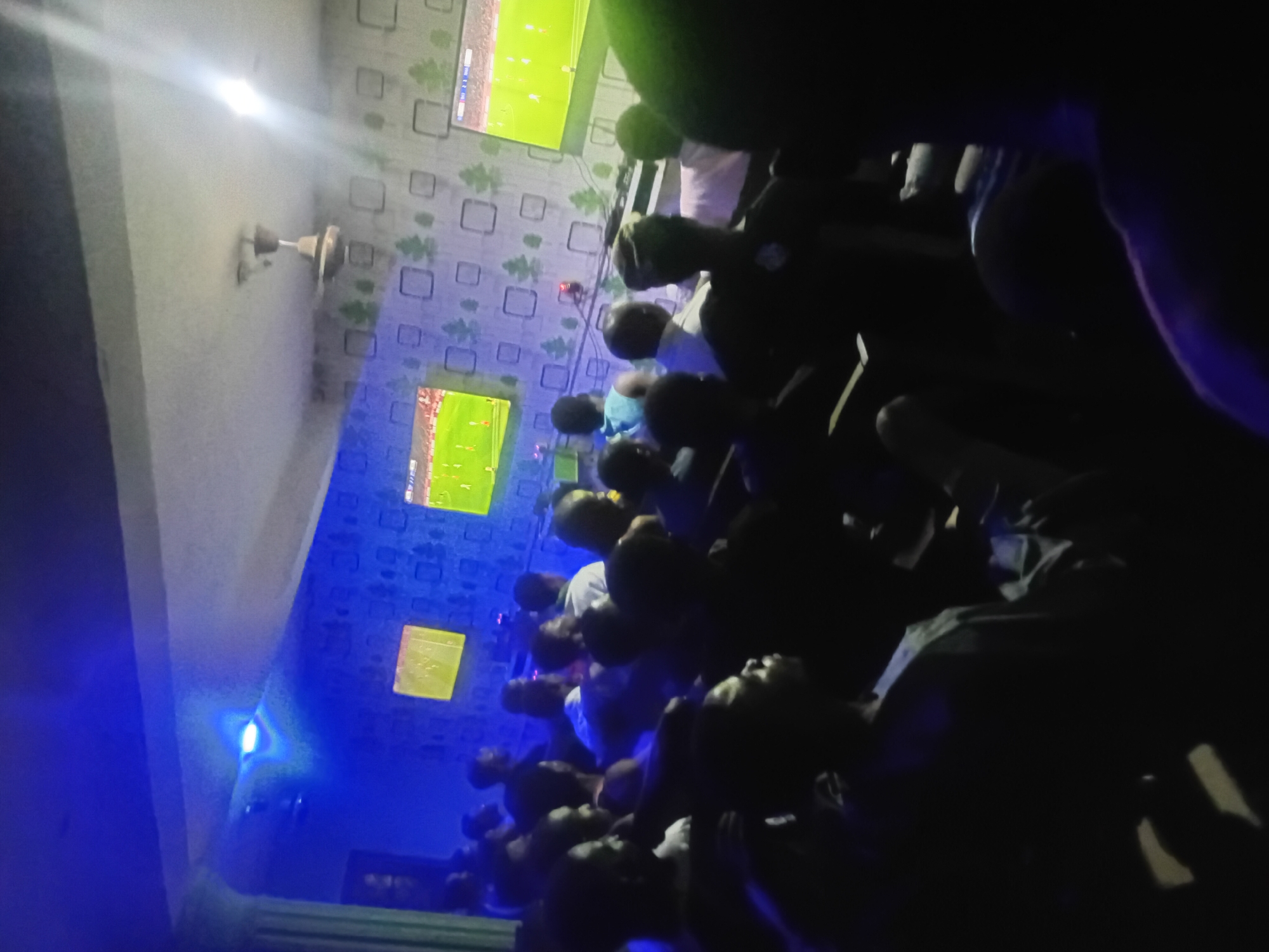 The image depicts a room in an unknown location with a crowd of people watching multiple televisions mounted on a wall. The wallpaper features a repeating square and floral pattern. The room is dimly lit with a blue hue. A ceiling fan is also visible.