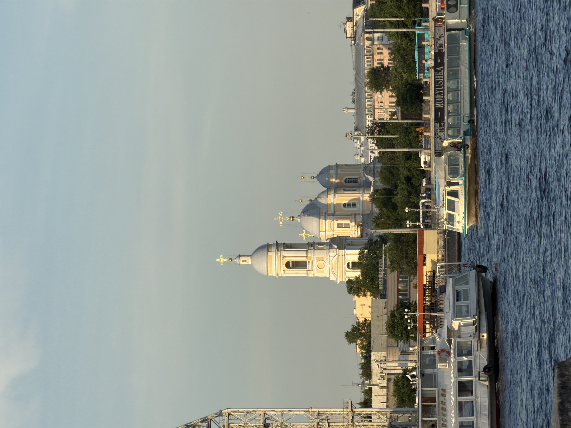 This is a full-frame, eye-level, vertical, outdoor shot of a riverfront scene in Saint Petersburg, Russia, under a clear, pale blue sky. The focal point is a body of water with two tour boats navigating it. The boat on the left is white with numerous windows, and the name "МОСКВА-64" is visible on its side. The boat on the right, also white, has the name "KORYUSHKA" prominently displayed in black lettering. Several people are visible on the decks of both boats.
In the background, a cluster of buildings with distinctive domed architecture, characteristic of Russian Orthodox churches, stands out. The largest structure features a tall, yellow bell tower topped with a golden cross. Adjacent to it are other domed buildings, also painted yellow. A tall, industrial-looking metal tower with a lattice structure is present on the far left of the image. Beyond the church complex, a line of urban buildings of various colors, including beige, pink, and light brown, is visible. Green trees punctuate the landscape between the river and the buildings. On the riverbank, a blue bus and several figures are discernible, along with a decorative metal railing. The water is dark blue and appears slightly rippled.
