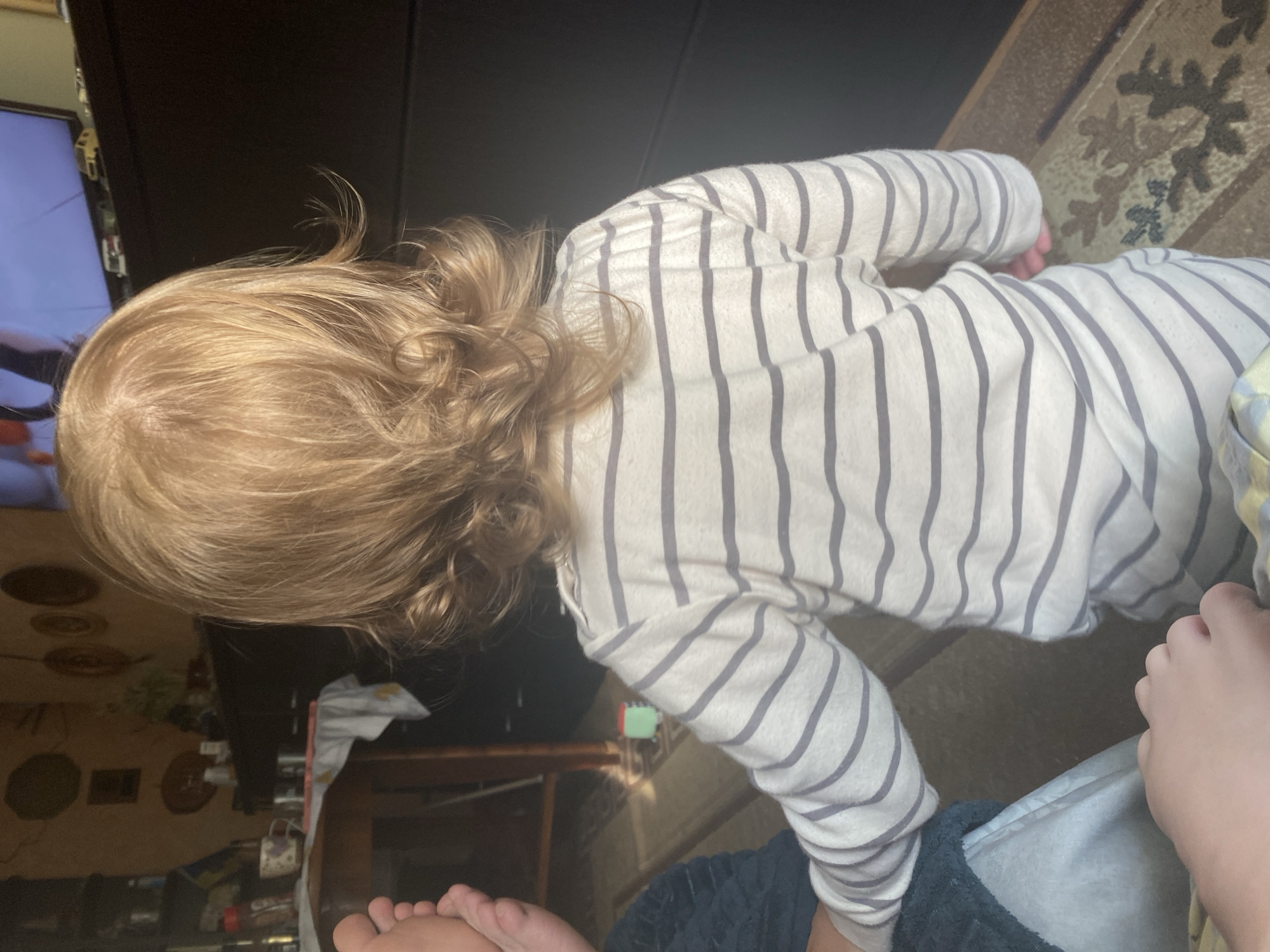 An overhead shot shows a child with light brown hair viewed from the back. The child is wearing a long-sleeved striped shirt. To the left, a person's foot is visible, and to the bottom, another person's hand rests on a blanket. The child is facing a dark surface and what appears to be a television screen showing a landscape. The background includes a table with items on it, and a wall with decorative plates. The floor is covered with a carpet.
