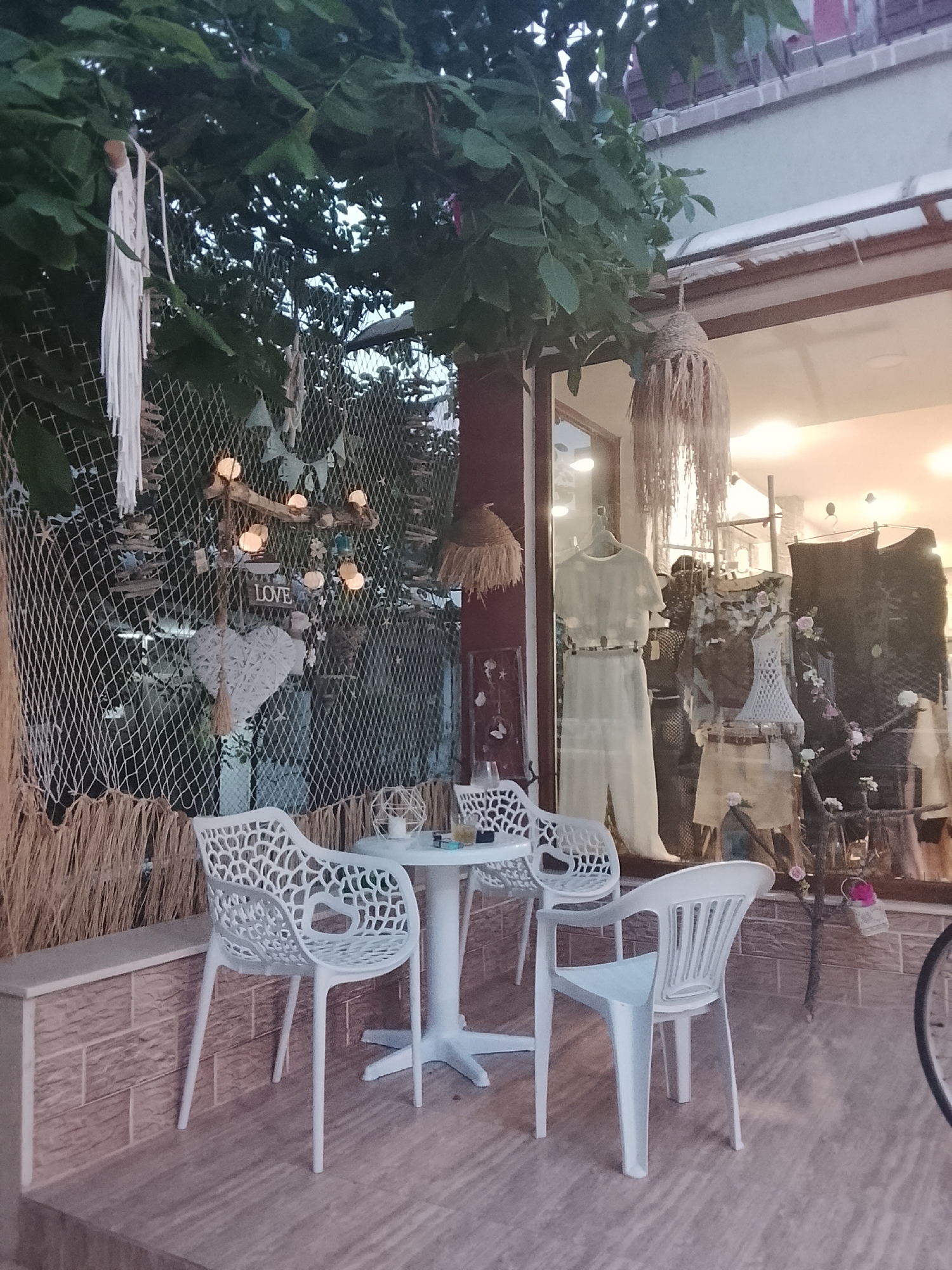 This image depicts a street-level view of a small shop's outdoor seating area, likely in Nesebar, Bulgaria, given the architectural style and coastal atmosphere. The scene is set at dusk, with the sky faintly visible through dense green foliage hanging above.
In the foreground, a white round table is set with three white, modern plastic chairs, two of which are visible in their entirety. The chairs have a distinctive openwork design. On the table, there is a white object, possibly a candle holder or a small decorative item, and a clear glass with some liquid and what appears to be a cigarette butt. A second table, partially obscured, is also visible.
Behind the seating area, a net-like fence is adorned with various decorations. These include a white macrame string decoration hanging on the left, strings of small, warm-toned fairy lights, and a prominent white heart-shaped decoration with tassels. A wooden sign with the word "LOVE" is also visible. Straw-like material is used as a border along the bottom of the fence and the exterior wall.
The shop window, reflecting the interior lights, showcases mannequins displaying clothing. One mannequin on the left is dressed in a cream-colored two-piece outfit. Another mannequin in the center wears a mesh top with a floral embellishment and a tan skirt. To the right, darker garments are displayed. The interior of the shop appears well-lit and decorated with natural materials, including straw-like hanging lamps. A small decorative tree with pink flowers is visible inside the window.
The ground is paved with textured tiles. The overall ambiance suggests a quaint, bohemian-style boutique or cafe. No people are directly visible in the frame.