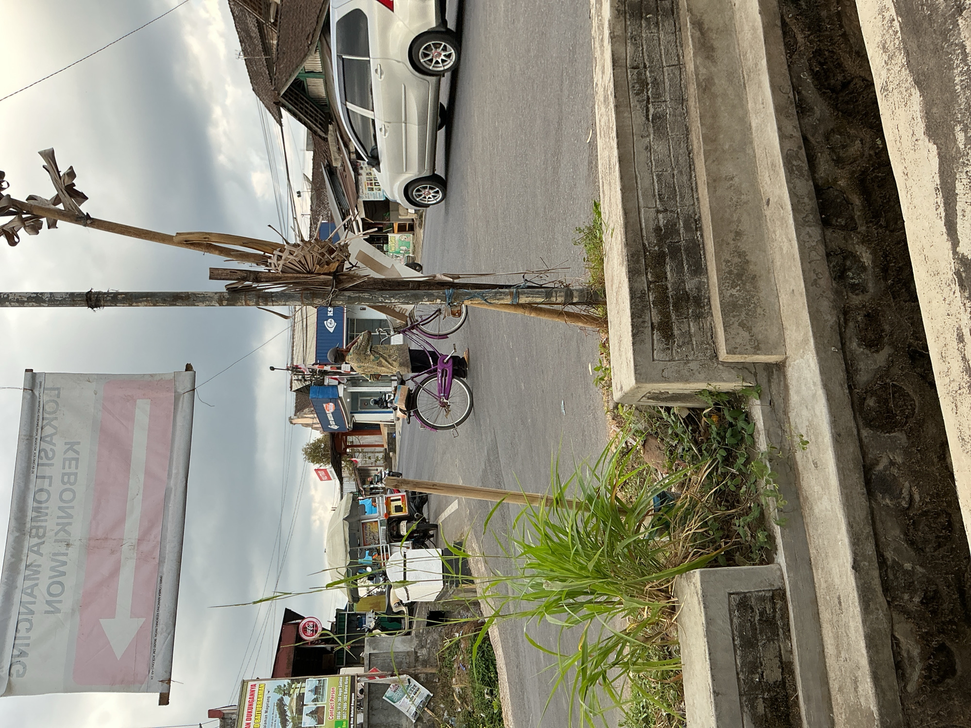 The image depicts a street scene in Kebonrejo, Indonesia, on an overcast day. In the foreground, concrete structures with visible brickwork suggest a drainage system or raised pathway. Lush green grass and weeds grow around these structures and on the side of the asphalt road. A wooden pole, adorned with dried leaves and bound with string, stands vertically on the roadside. A person wearing a patterned shirt and dark pants is seen standing next to a purple bicycle, which has a bag or box attached to its rear rack. Further down the road, a silver minivan is in motion, and other vehicles, including a three-wheeled cart and motorcycles, are visible in the background. Buildings line the street, with some featuring tiled roofs and signage. A large banner, hanging across the street, displays text in a language that appears to be Indonesian, with a prominent arrow pointing to the left. The overall atmosphere is that of a typical urban environment during a cloudy day.