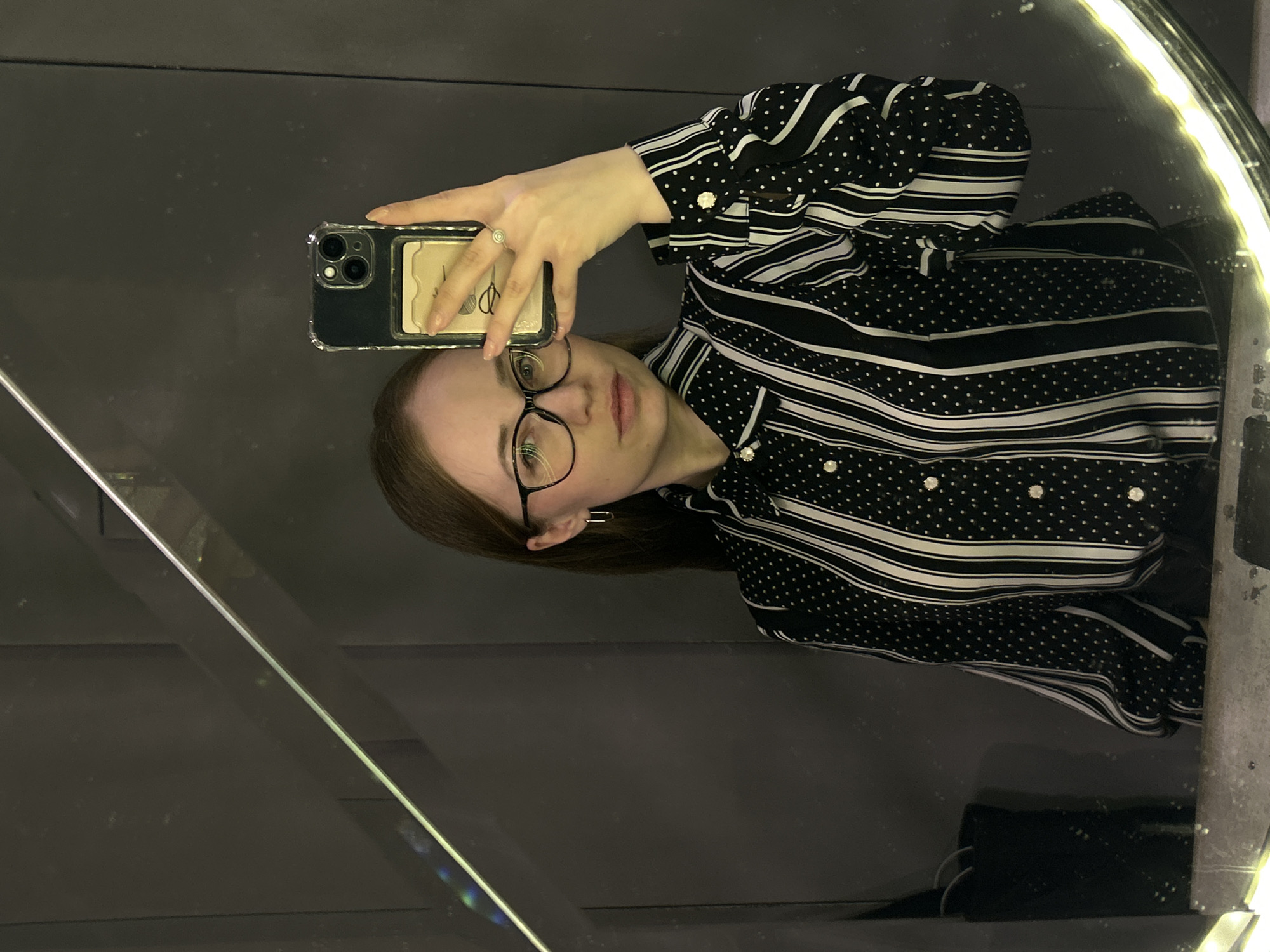 A mirror selfie captures a young woman in an indoor setting, identified as Saint Petersburg, Russia. She has shoulder-length light brown hair and wears black-framed glasses. Her expression is neutral, and her gaze is directed towards the reflected smartphone camera. She is dressed in a black and white patterned long-sleeved shirt, featuring sections with thin white horizontal stripes and other sections with small white polka dots.
In her right hand, she holds a dark-colored smartphone, likely an iPhone model with a multi-lens camera system, encased in a clear protective cover. A light-colored card with partially obscured text, possibly resembling a brand name like 'LOEWE', is visible tucked into the back of the phone case. A silver ring adorns her finger.
The environment reflected in the mirror appears dark and glossy, suggesting dark walls or panels. A prominent bright, circular light, characteristic of a ring light, is reflected around the upper right edge of the mirror, providing illumination. A diagonal reflective strip or edge is visible in the lower left of the frame. The scene conveys an intimate, perhaps private, moment of self-photography within an interior space, illuminated by artificial light.