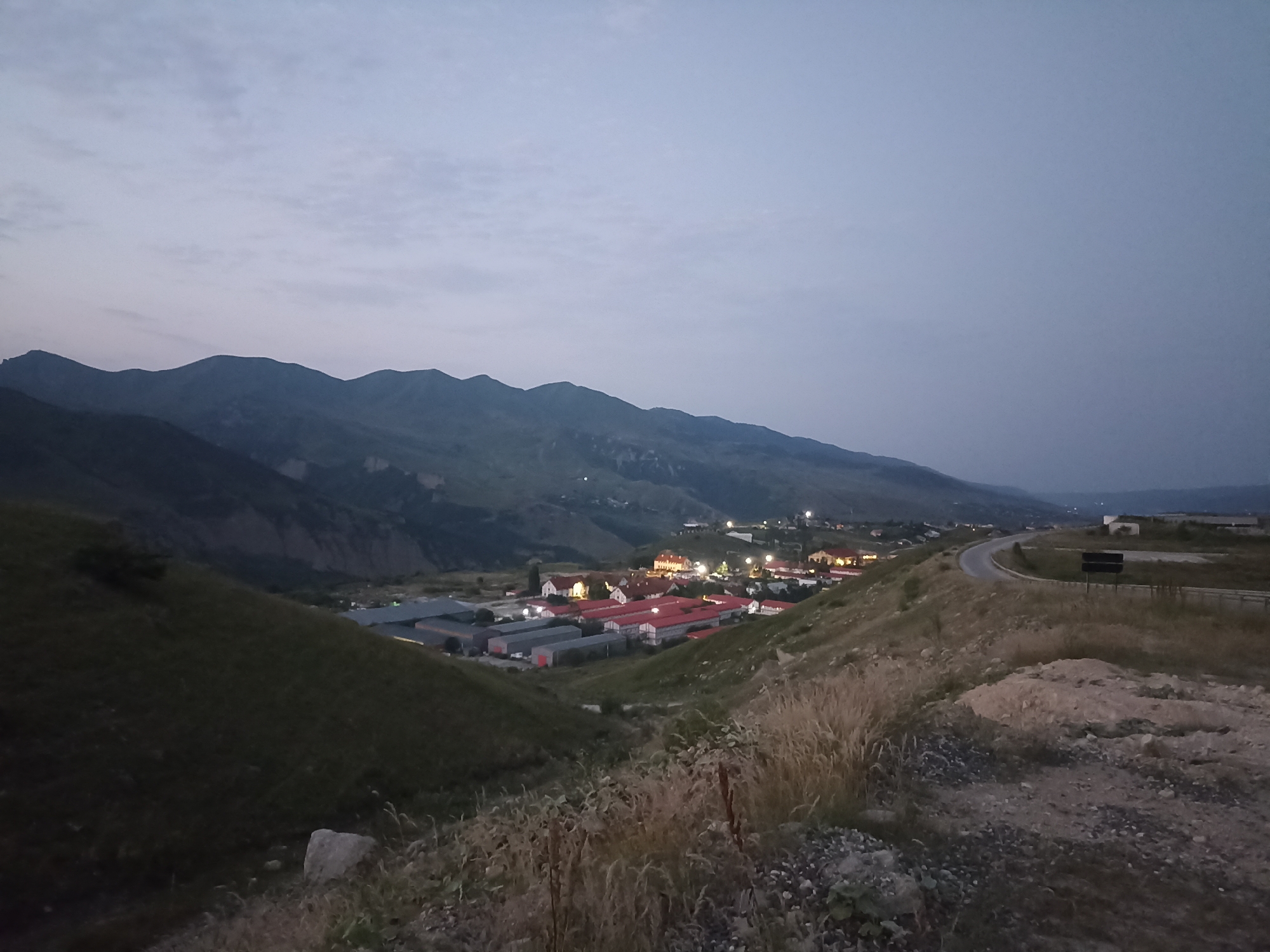 The image depicts a twilight view of a mountainous landscape with a settlement in the valley below, identified as Kuzun, Azerbaijan. The sky is a pale, overcast blue transitioning to darker hues near the horizon. In the foreground, a grassy, sloped terrain is visible, with dry grasses and rocks scattered across it. A dirt road or path runs along the upper right edge of the foreground.
In the mid-ground, a collection of buildings forms a village or small town. These structures have red tiled roofs and appear to be illuminated by interior lights, suggesting it is early evening. Some of these buildings are larger, industrial-looking structures with dark grey roofs. A paved road curves through the settlement and continues into the distance.
The background is dominated by large, dark, undulating mountains that recede into the haze. The overall atmosphere is serene and quiet, with no people or animals visible in the frame. The lighting suggests a time of day shortly after sunset, with artificial lights from the settlement becoming prominent against the fading natural light.