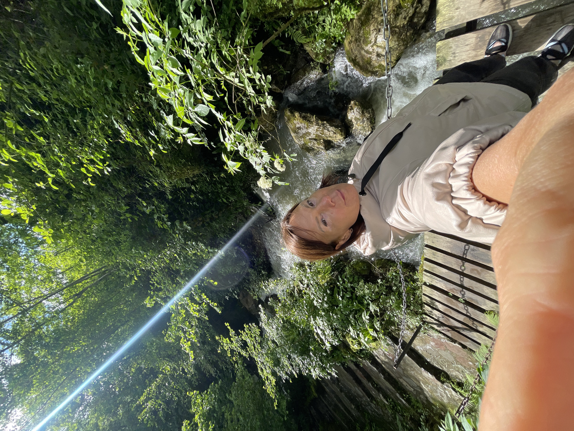 A woman stands on a wooden bridge above a small, fast-flowing stream, possibly a minor waterfall, in a densely vegetated area. The location is in Solokh-Aul, Russia. The environment is characterized by lush green trees and vegetation. The woman wears a light-colored jacket, dark pants, and shoes. She looks directly at the camera, seemingly taking a selfie. The photo captures the scene from an angled perspective. A long chain hangs from the bridge.