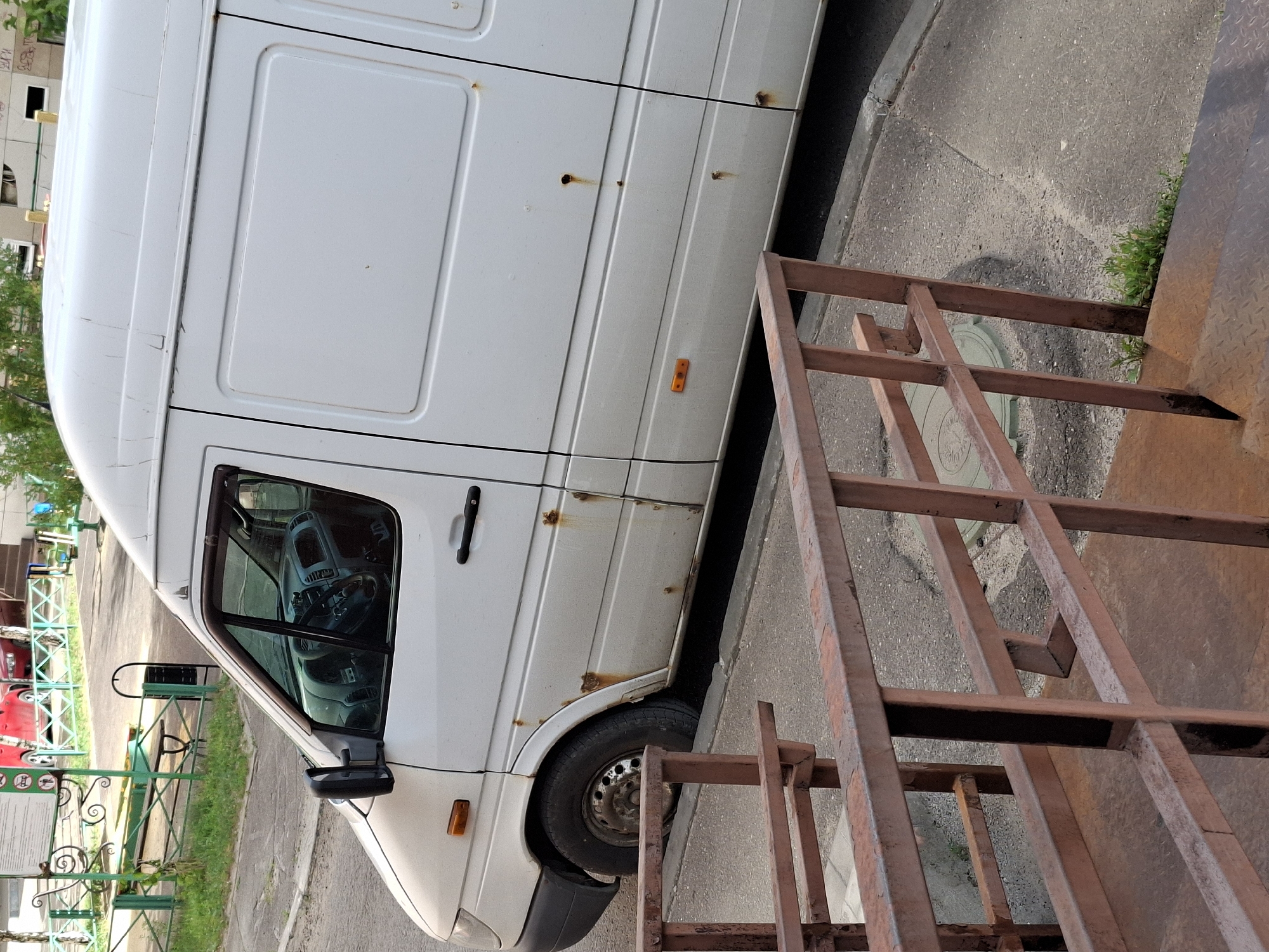 The image shows the side of a white cargo van parked on an asphalt surface. The van has visible rust spots and some dents. A metal staircase, appearing to be made of rusted metal bars, is positioned to the right of the van, partially obscuring the side of the vehicle. The staircase is angled, suggesting it might be a temporary structure or part of a larger construction.
In the background, a building with multiple windows is visible, along with some trees and greenery. A green metal fence with decorative elements and some signage is also present. The overall scene appears to be an outdoor urban or semi-urban environment, possibly a residential area or a service area. The lighting suggests it is daytime, with no specific indication of the time of day or weather conditions. There is no visible activity or people in the frame. The crops provide closer views of the van's body, the staircase, and the surrounding environment.