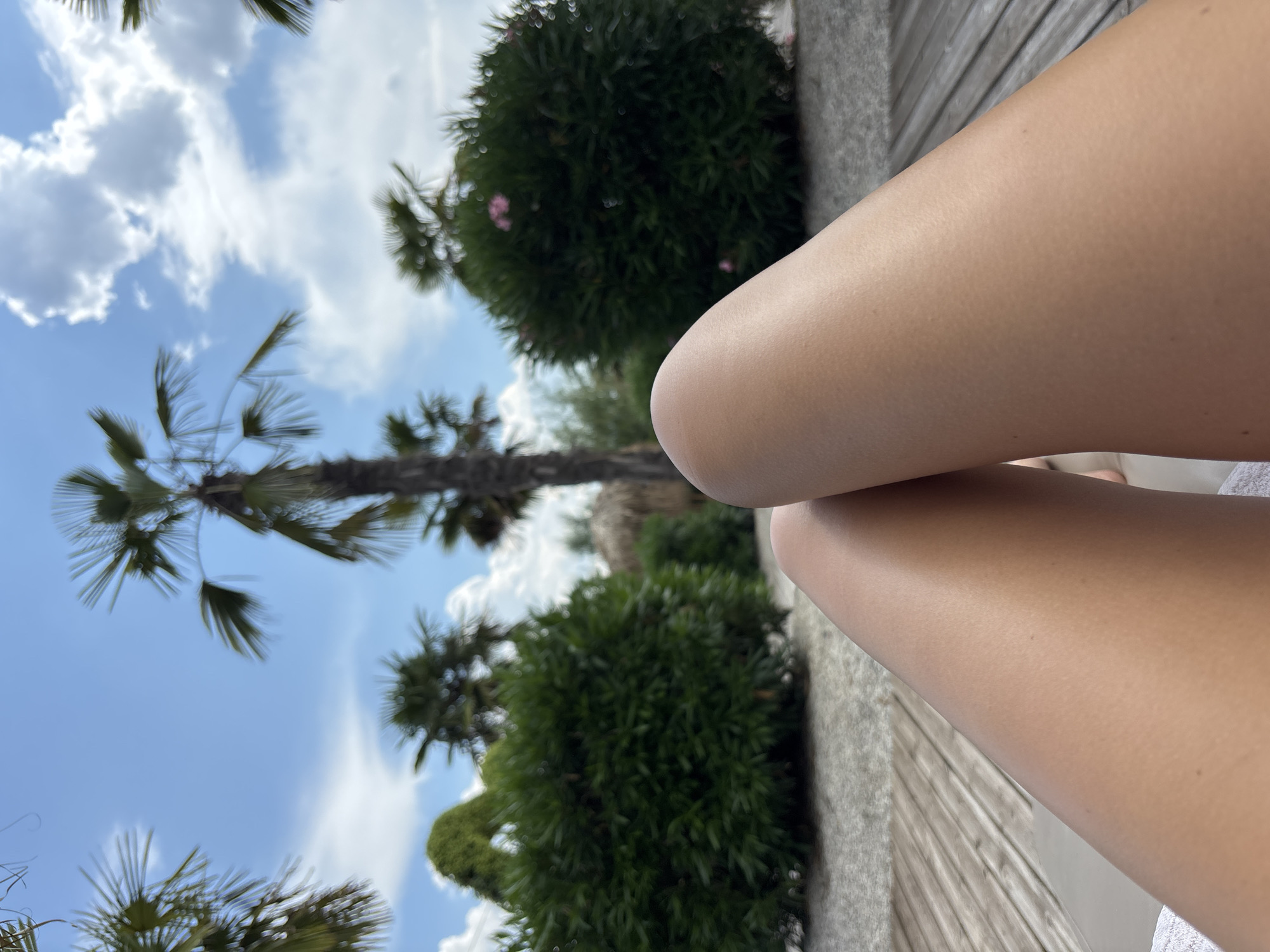 The image displays a close-up view of a person's bare legs resting on a wooden deck. In the background, lush green bushes and palm trees are visible under a bright blue sky with scattered white clouds. A small thatched-roof structure can be seen between the foliage. The overall scene suggests a tropical or resort-like setting, possibly a poolside area or a garden lounge. The specific location in Plovdiv, Bulgaria, cannot be determined from the visual information provided.