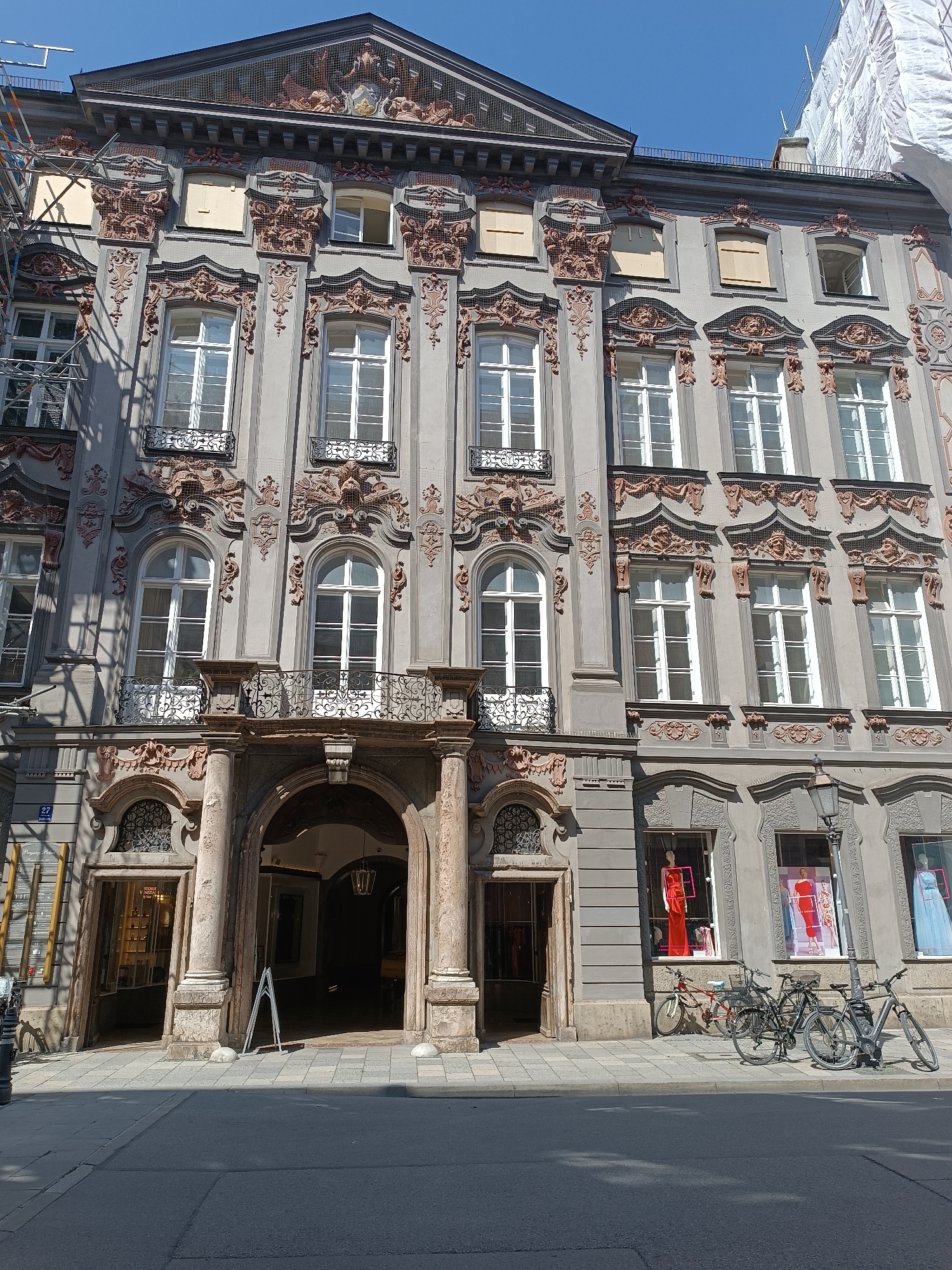 The image shows a historic building in Munich, Germany, with ornate architecture and arched doorways. The building appears to be a shop or commercial establishment on the ground floor, with several windows displaying mannequins dressed in formal attire. Several bicycles are parked in front of the shop, suggesting a street-level commercial area. The overall impression is of an urban setting with a blend of historical and modern elements. The building's facade is detailed with decorative elements and multiple windows, some of which are covered or boarded up, indicating possible ongoing renovations or maintenance. The street in front of the building is paved, and shadows from the building and trees are cast across it. The sky is clear and blue, suggesting a sunny day. The specific location within Munich is not provided in the image.