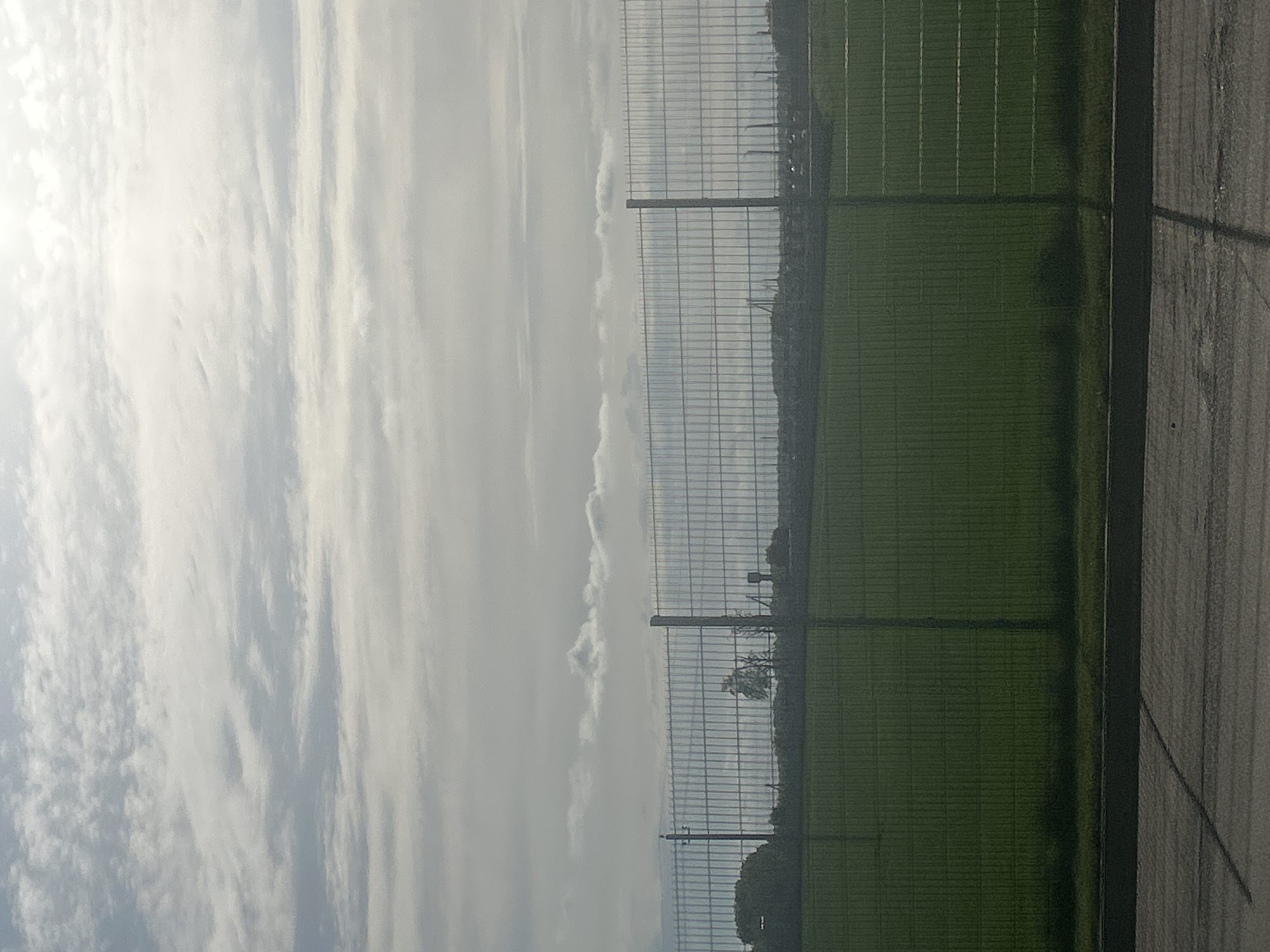The image shows a section of what appears to be a sports field or park in Міханавіцкі сельскі Савет, Belarus, based on the user's location. On the right, there's an artificial turf field, segmented with white lines. A dark border separates the field from a gray, possibly paved, area. A chain-link fence runs along the field's edge, partially obscuring the view of trees in the background. The sky is overcast, with a predominance of light gray and white clouds. There are no people visible in the image.