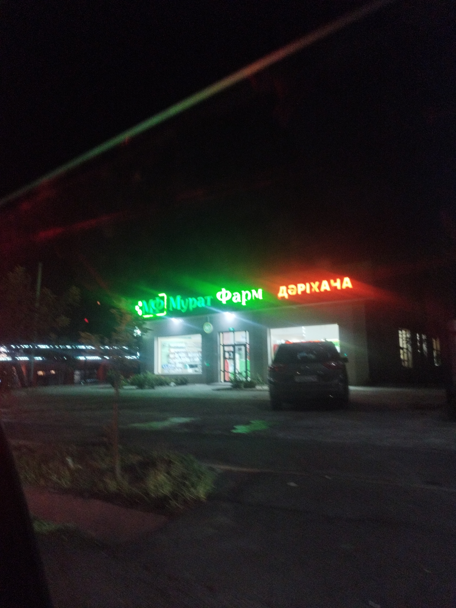 The image shows a nighttime street scene in Shymkent, Kazakhstan. A brightly lit sign above a building reads "MURAT ФАРМ ДӘРІХАНА" in green and orange neon letters, indicating a pharmacy. A dark-colored SUV is parked in front of the pharmacy entrance. The building appears to be a commercial establishment with glass windows. Trees and vegetation are visible to the left of the building. The street and surrounding area are dark, with the primary illumination coming from the building's signage and interior lights.