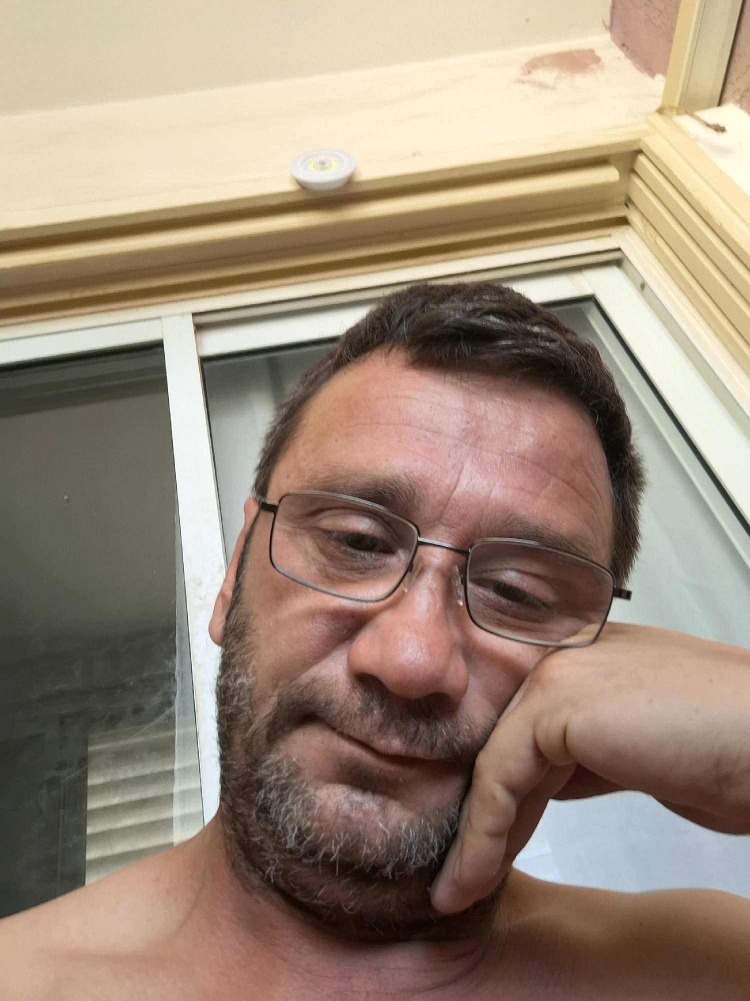 The image is a close-up selfie of a man in Yerocham, Israel. The man has short dark hair, glasses, and a goatee. He is resting his head on his hand, looking downwards and slightly to his left. The background shows a window and a light fixture mounted on a wooden trim. The lighting suggests it is daytime.