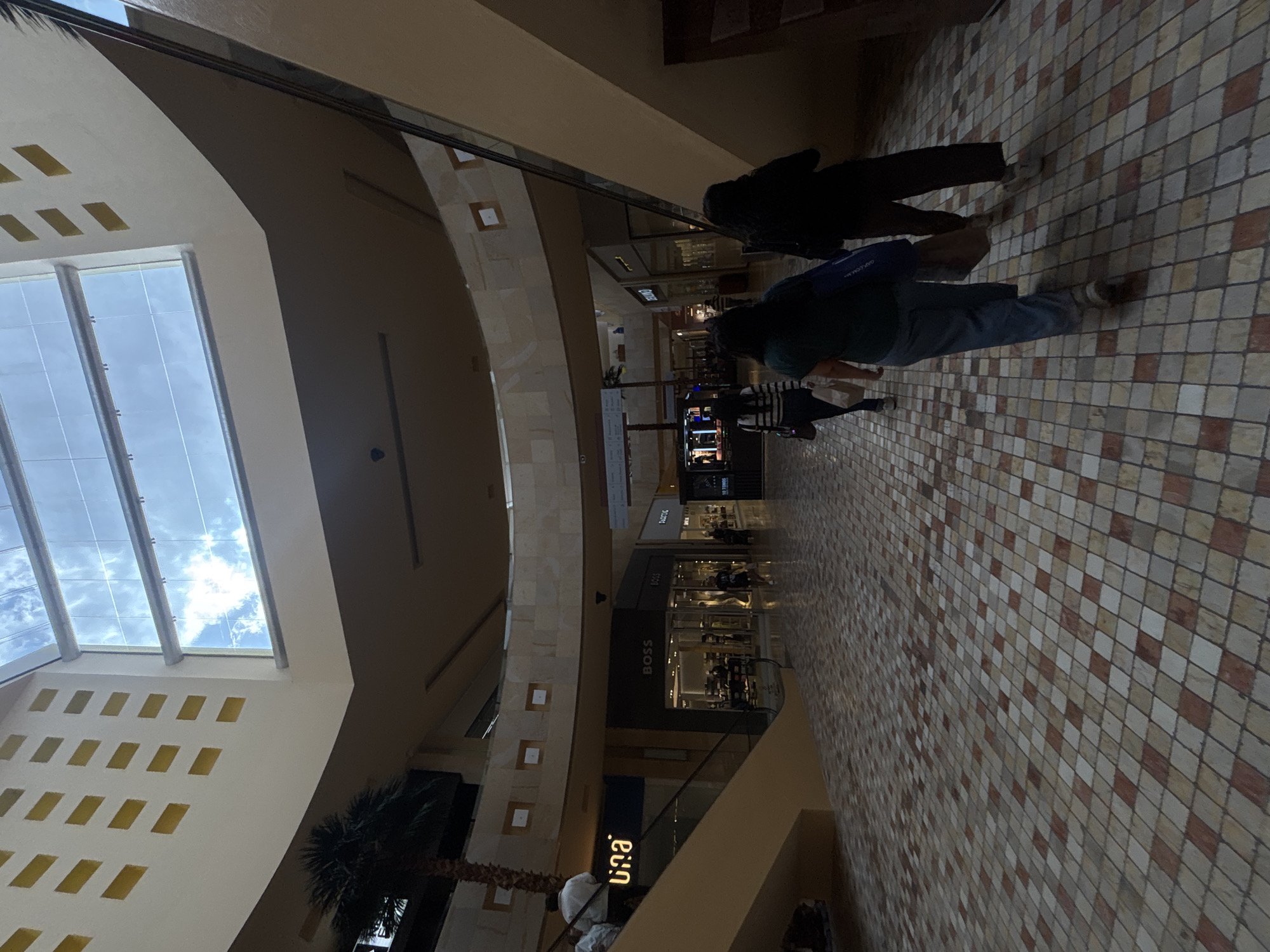The image shows the interior of what appears to be a shopping mall, likely located in Puebla, Mexico, given the provided context. The architecture features a large skylight, allowing natural light to illuminate the indoor space. The interior walls are decorated with a geometric pattern. A tall palm tree is visible near the base of the skylight. Several stores are visible along the mall's corridors, including "BOSS" and "una" brands. The floor is tiled in a checkered pattern of light and dark squares. Several people are walking through the mall. Two people are walking closer to the camera.