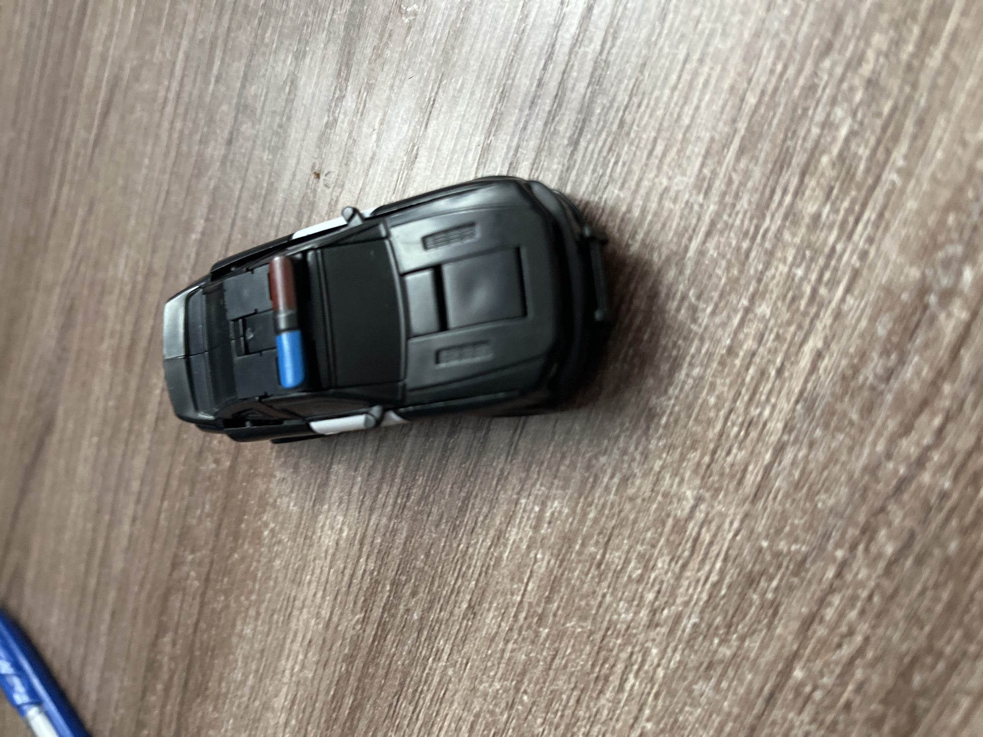 A toy car rests on a wood-grain surface. The car is black, with what appears to be a police light bar on top in red and blue. A blue pen is partly visible at the lower left corner.