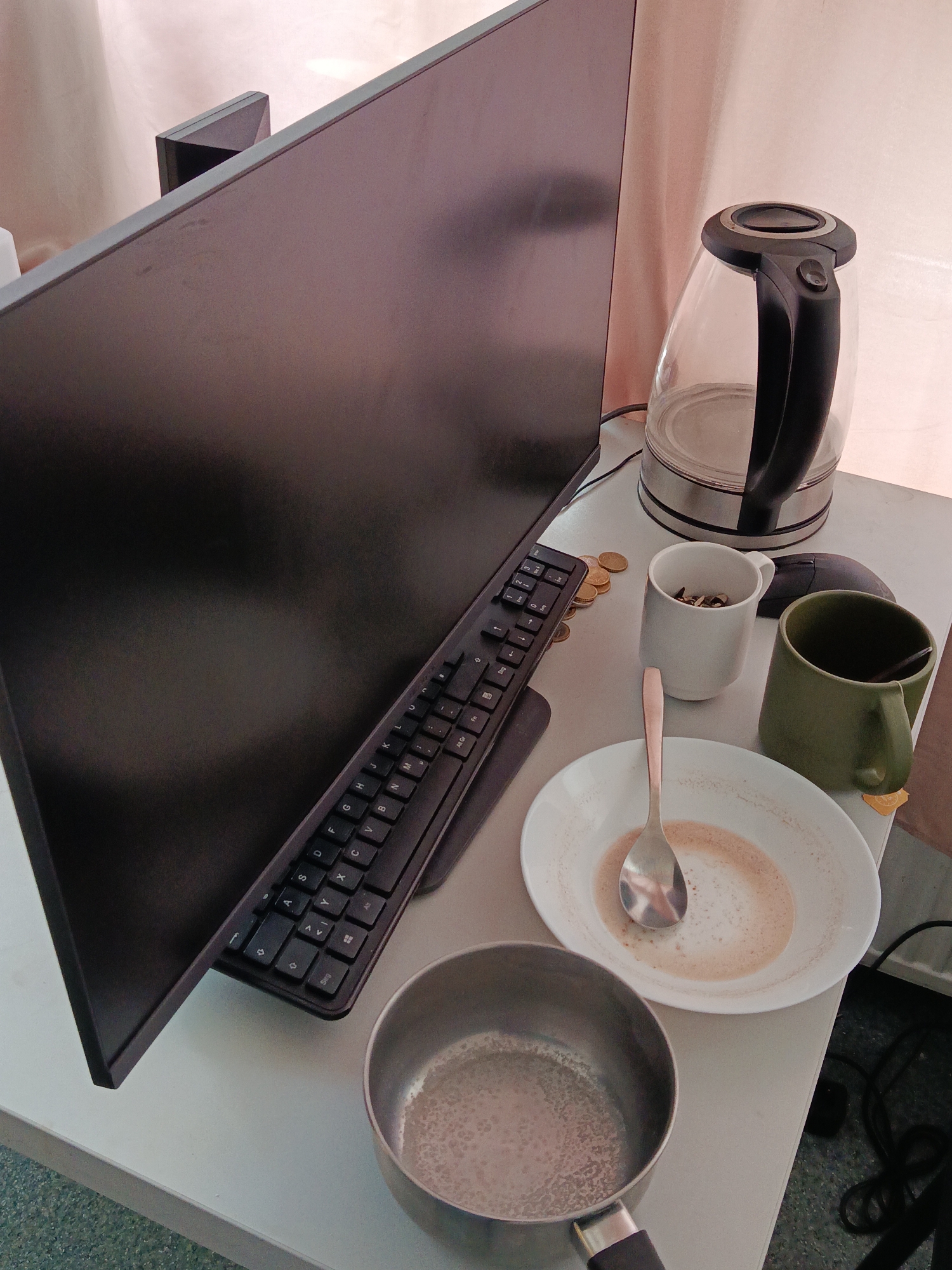 The image depicts a desk with a computer monitor, keyboard, mouse, and various items related to drinking coffee or tea. A transparent electric kettle is situated to the right of the monitor. In front of the monitor, a keyboard is visible, and to its right, a stack of coins, a white mug with a dark substance, a green mug with a spoon, and a white bowl with residue and a spoon are arranged. A small metal pot with a handle is also present. The scene appears to be indoors, on a desk with a light-colored surface, and a blurred light-colored curtain is visible in the background. There are no people or animals in the image, and no specific location in Halbe, Germany, can be identified from the visual cues.