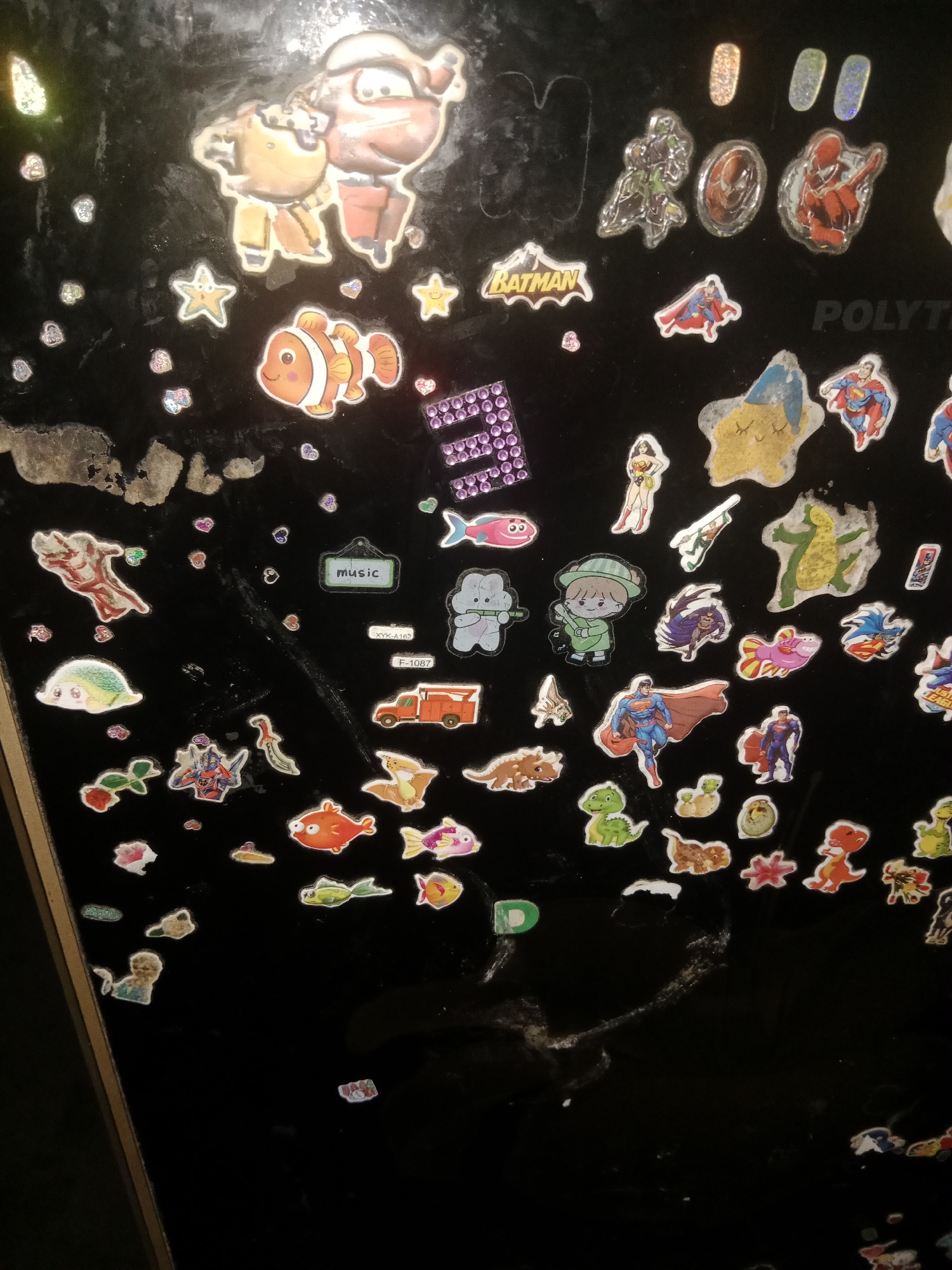 A black, glossy surface, likely a refrigerator door, is covered in a variety of colorful stickers. The stickers depict cartoon characters, including a clownfish, superheroes like Batman and Superman, various dinosaurs, and other whimsical figures such as stars and hearts. In the upper right portion of the surface, the word "POLYT" is visible, possibly part of a logo or label. The surface itself shows some wear and tear, with scuffs and marks. The image is a close-up shot, with no visible background or surrounding environment.