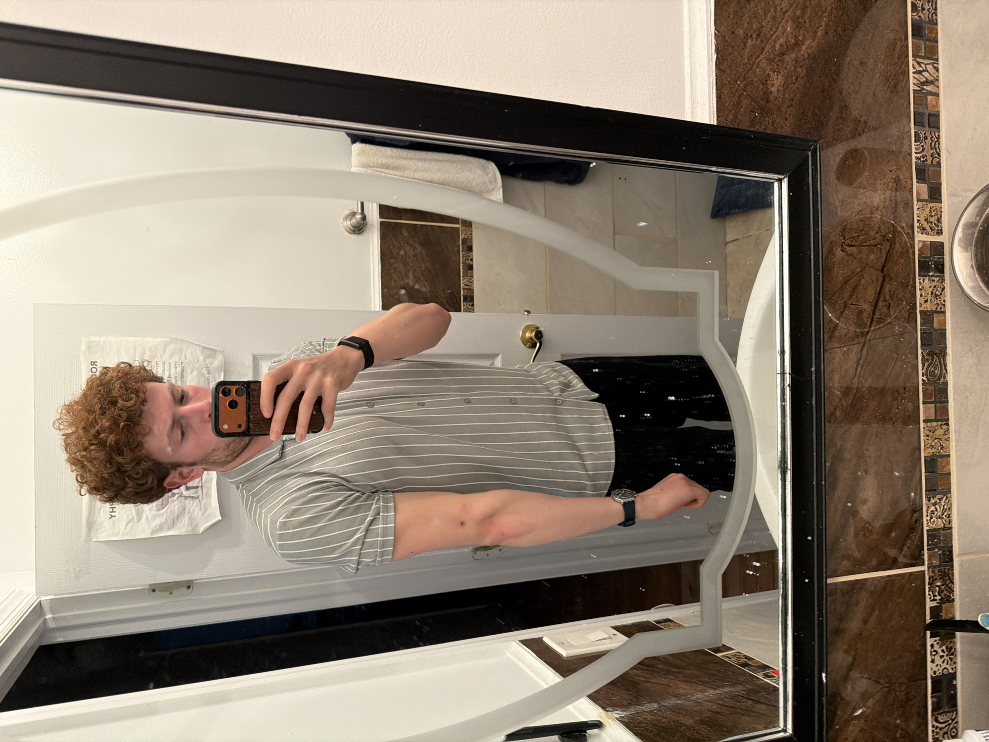A reflection in a bathroom mirror shows a man taking a selfie. The man has curly brown hair and is wearing a light gray striped shirt. He is holding a phone with a wood-grain case. He is also wearing a smartwatch on his left wrist and a wristwatch on his right wrist.
The bathroom features a white door with a paper taped to it, a white sink, and brown and tan tiled walls. A shower curtain rod is visible above the shower. A white towel is on the shower curtain rod.