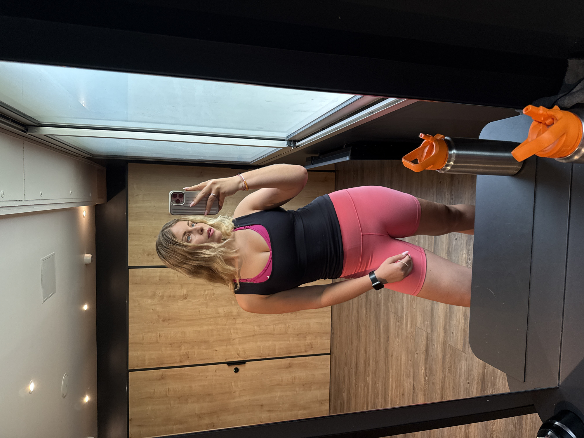 A blonde woman is standing in front of a mirror, taking a selfie. She is wearing a black tank top over a pink sports bra and pink biker shorts. She has a black watch on her left wrist and a ring on her right ring finger. Her hair is wavy and falls to her shoulders. The background appears to be a gym or fitness studio, with wooden walls and a large window. There is a metal water bottle with an orange lid on a black surface in the foreground, and another similar bottle partially visible. The overall scene is well-lit. There is no indication that this scene is located in Paris, France.