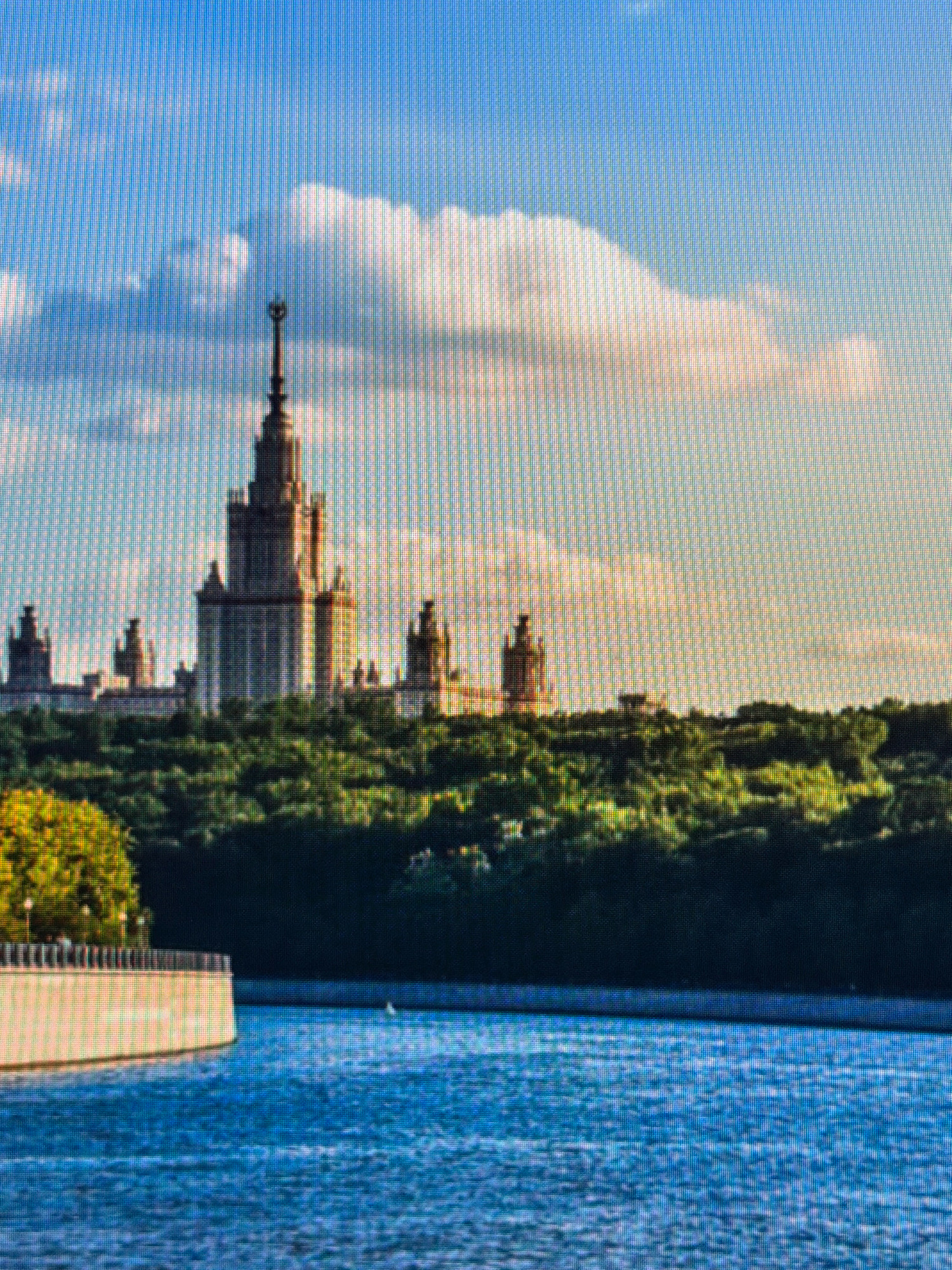 The image depicts a vast body of water in the foreground, reflecting the blue sky and scattered clouds. A curved concrete embankment, lined with a railing, runs along the left side of the water. Beyond the embankment, a dense green forest stretches across the middle ground. A grand, multi-story building with a central spire and several smaller adjacent structures rises above the trees in the background, silhouetted against the sky. The sky itself is a clear blue, with a large, fluffy white cloud dominating the upper portion of the frame, tinged with a warm, golden light from what appears to be a sunset or sunrise. The overall scene is serene and picturesque, evoking a sense of grandeur and natural beauty. The location is not Kafr Yasif, Israel.