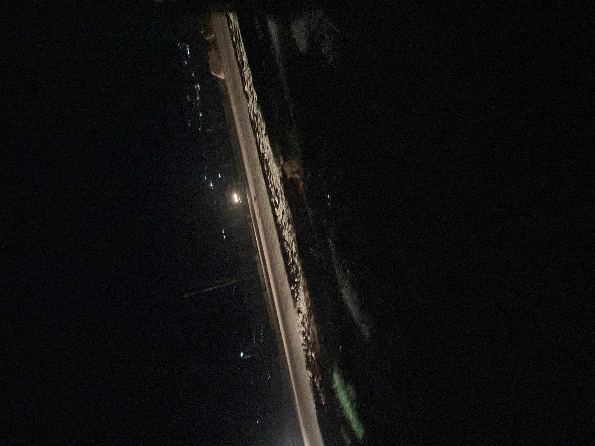 The image depicts a dark, nighttime scene on what appears to be a road in Lokoja, Nigeria. The road itself is visible, with a lighter-colored surface, possibly asphalt or gravel, illuminated by an unseen light source. To the right of the road, there is a rough, uneven embankment with patches of greenery and scattered debris. In the distance, along the horizon, a few scattered lights indicate the presence of buildings or streetlights. The overall impression is one of a somewhat undeveloped or rural area within or on the outskirts of the city, characterized by low light conditions and a natural, unmanicured environment. There are no people or vehicles clearly identifiable in the image.