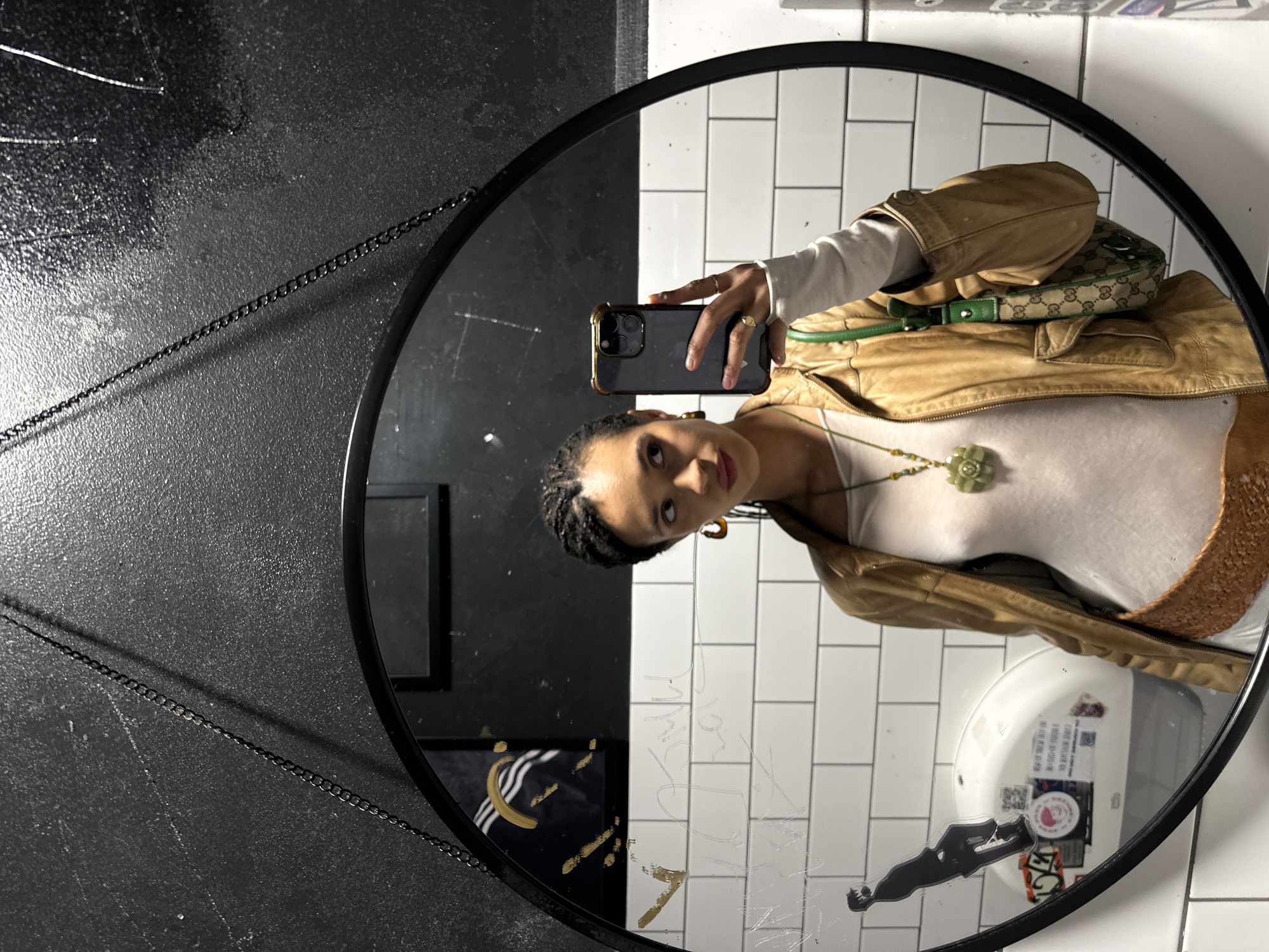 A person is captured in a mirror selfie, standing upright, reflected in a round, black-framed mirror. The mirror is hung by chains against a dark, textured wall. The individual is a female with dark skin and braided hair, wearing a white long-sleeve top under a light brown, possibly leather, jacket. A white tank top is visible beneath the jacket, along with a long green necklace featuring a floral pendant. She has gold-toned earrings and rings on her fingers. She holds a dark-colored smartphone with a gold-rimmed case in her right hand, pointing it towards the mirror to take the photo, looking directly into the camera.
A small Gucci handbag with a green strap and the brand's signature pattern is slung over her left shoulder, also reflected in the mirror. The background visible within the mirror's reflection consists of a white-tiled wall, suggesting an indoor setting like a restroom. Various faint scratches and what appears to be etched or drawn graffiti are visible on the mirror's surface, including some gold-colored marks, one resembling "YA" or "VA" at the bottom. Reflected in the bottom right, a sink and faucet are partially visible, covered with stickers, some bearing QR codes and small, illegible text. To the left within the mirror's reflection, part of a dark framed object with a gold Adidas logo (three stripes and a semicircle) is visible. The scene is brightly lit by indoor lighting.