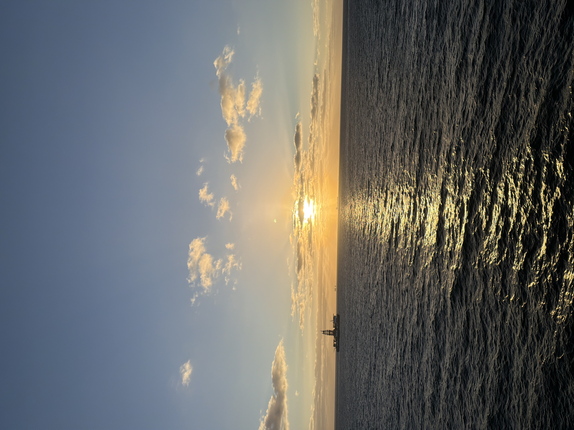 A landscape view shows a large body of water on the right side of the frame, reflecting sunlight. The sky occupies the left side, with a blue gradient transitioning to lighter hues near the horizon, where the sun appears. Clouds are scattered throughout the sky. A distant structure, possibly a ship or platform, is visible at the horizon line.