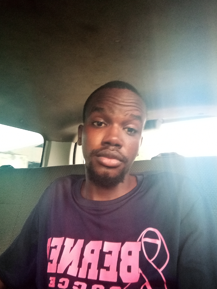 A dark-skinned male, with short dark hair and a beard, is depicted seated inside a vehicle. He is observed from the chest up, maintaining direct eye contact with the camera. He wears a dark-colored short-sleeved t-shirt featuring pink, reversed text that reads "BERNEE" or a similar variant, accompanied by a pink ribbon symbol on the left side of his chest (viewer's right). The vehicle's interior shows a light-colored headliner above and dark, textured seat upholstery behind the individual. Bright, diffused daylight is visible through the windows on both sides, indicating an exterior environment, but specific identifying features of Mtopanga ward, Kenya, are not discernible from the image.
