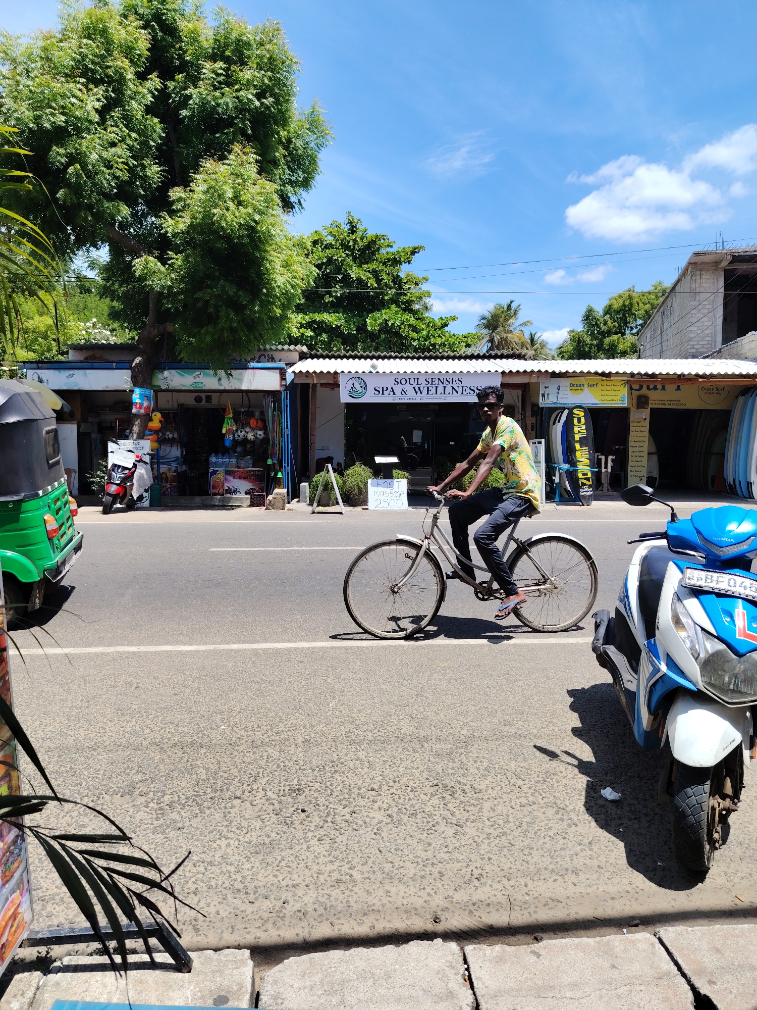 In an outdoor urban scene, a young man is cycling on a paved road. He is wearing a colorful t-shirt and dark pants, with sunglasses on his face. The man is mid-action, pedaling his bicycle. The background features several shops with awnings, displaying various items like surfboards. A bright green tuk-tuk is parked to the left of the road, and a blue and white scooter is parked to the right, partially obscuring the view of the road. The environment suggests a sunny day with a clear blue sky and some scattered clouds. Lush green trees are visible above the shops, contributing to the overall outdoor setting.
