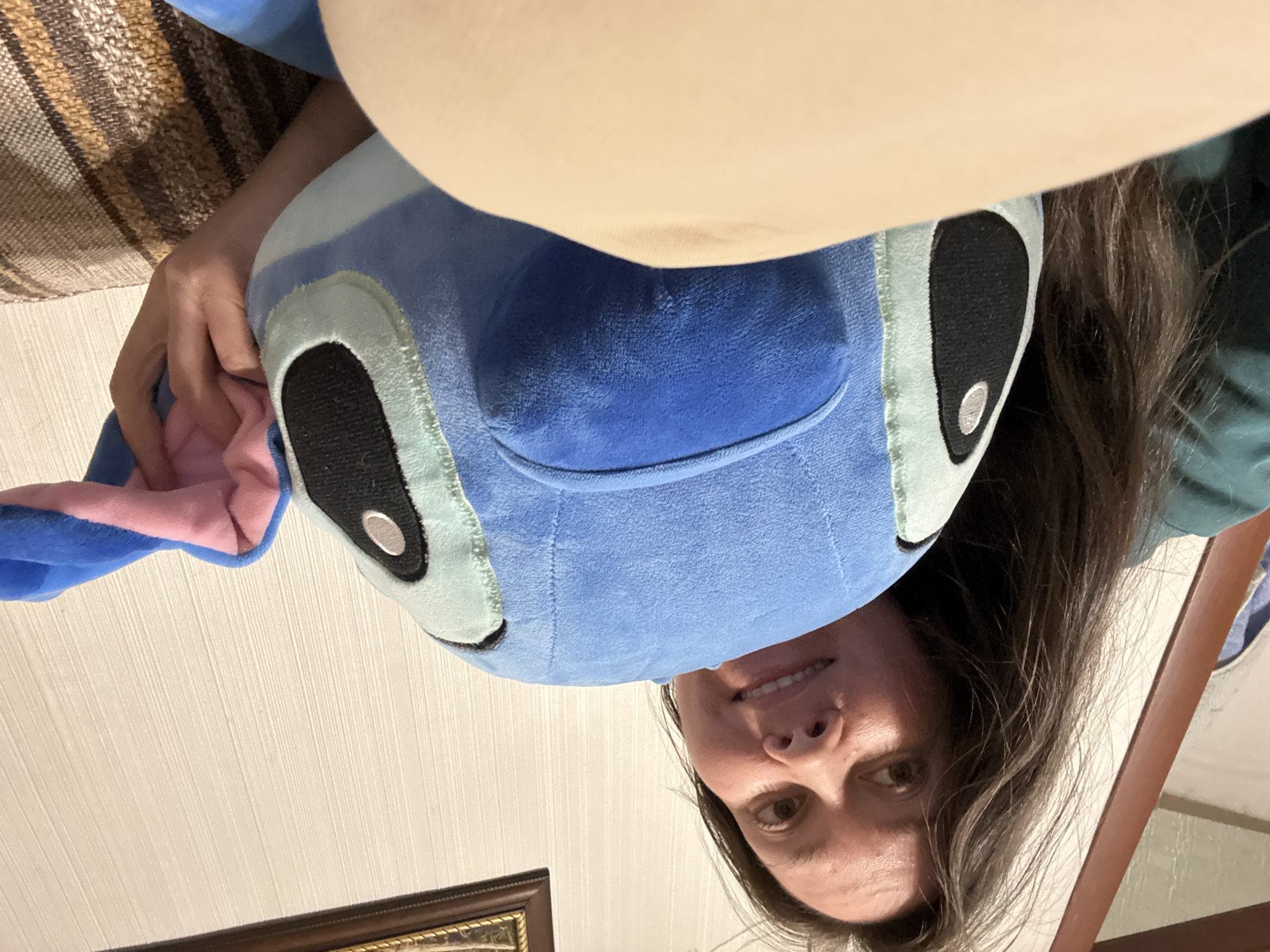 A person is holding a blue and pink plush toy with large black eyes. The person's face is partially visible below the toy. The location appears to be indoors, with a patterned wall and a picture frame visible in the background. A portion of a couch is visible. The location is not verifiable as Izhevsk, Russia based on the image content.