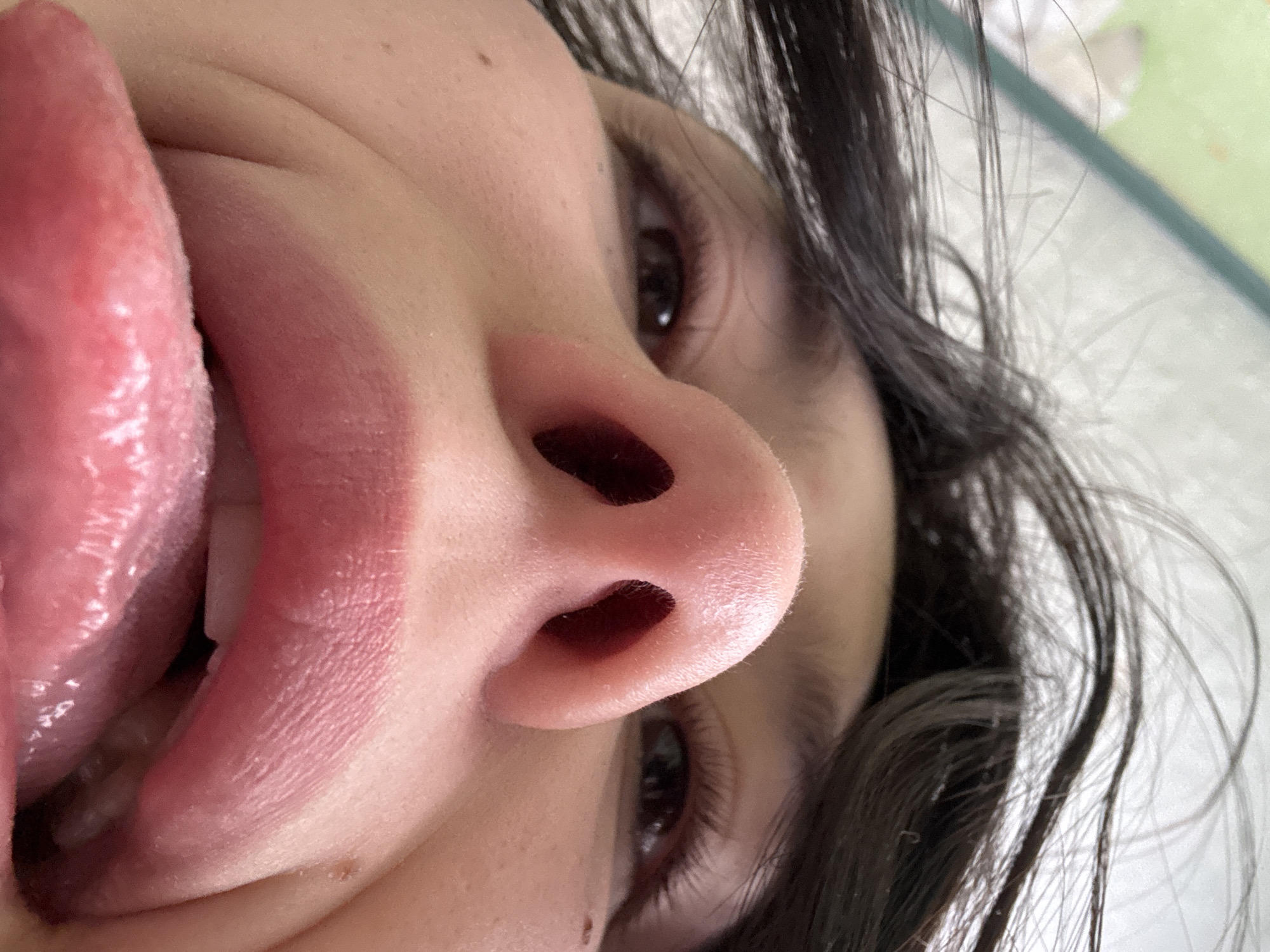 An extreme low-angle, close-up photograph captures the central facial features of an individual. The camera is positioned directly beneath the nose, which dominates the upper-middle frame, prominently displaying the nostrils. Dark hair is visible along the top edge of the image, framing the upper portion of the face. Below the nose, the subject's mouth is open, with full, pink lips from which a wet, pink tongue is extended downward and outward. A portion of the upper teeth is visible behind the tongue. Both eyes, possessing dark irises and visible eyelashes, are partially visible in the upper-left and upper-right quadrants, looking forward. The skin exhibits minor textural details. The background is out of focus and indistinct, showing faint light green and white tones, suggesting an indoor environment. The specific location within the city of Фоки, Russia, cannot be determined from the visual evidence presented in the image.