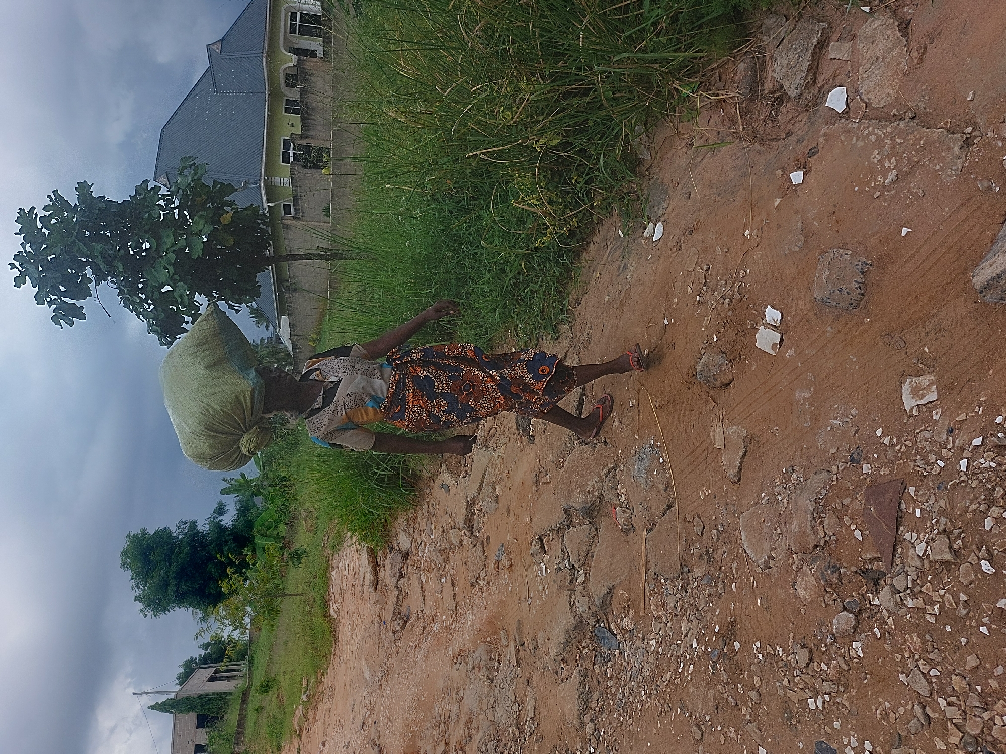 A woman carries a large, sack-like object on her head, walking along a rough, dirt path in an area with scattered debris and vegetation. A building is visible in the background. The location is likely in Dar es-Salaam, Tanzania.