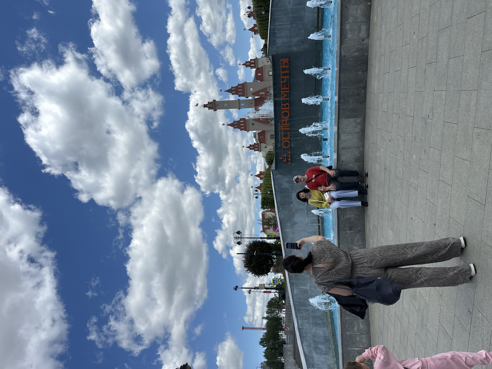 An exterior, eye-level, full shot shows a man and two women posing in front of a fountain with a castle-like structure in the background, under a bright blue sky filled with white, fluffy clouds. The castle in the background features multiple towers with red roofs and flags, and the words "ОСТРОВ МЕЧТЫ" (Dream Island) are visible in large orange lettering on a dark gray wall in front of it. Water jets from the fountain spray upwards into the air.
In the foreground, a woman wearing a patterned jumpsuit is holding up a smartphone, appearing to take a picture. To her right, a couple is seated on the edge of the fountain. The woman wears a yellow top and a white purse, and the man wears a red t-shirt. To the far left, a child in pink clothing is partially visible, with their back to the camera. The ground is paved with grey tiles. The overall setting appears to be an amusement park or entertainment complex, likely the "Dream Island" (Остров Мечты) amusement park in Moscow, Russia, given the prominent signage.