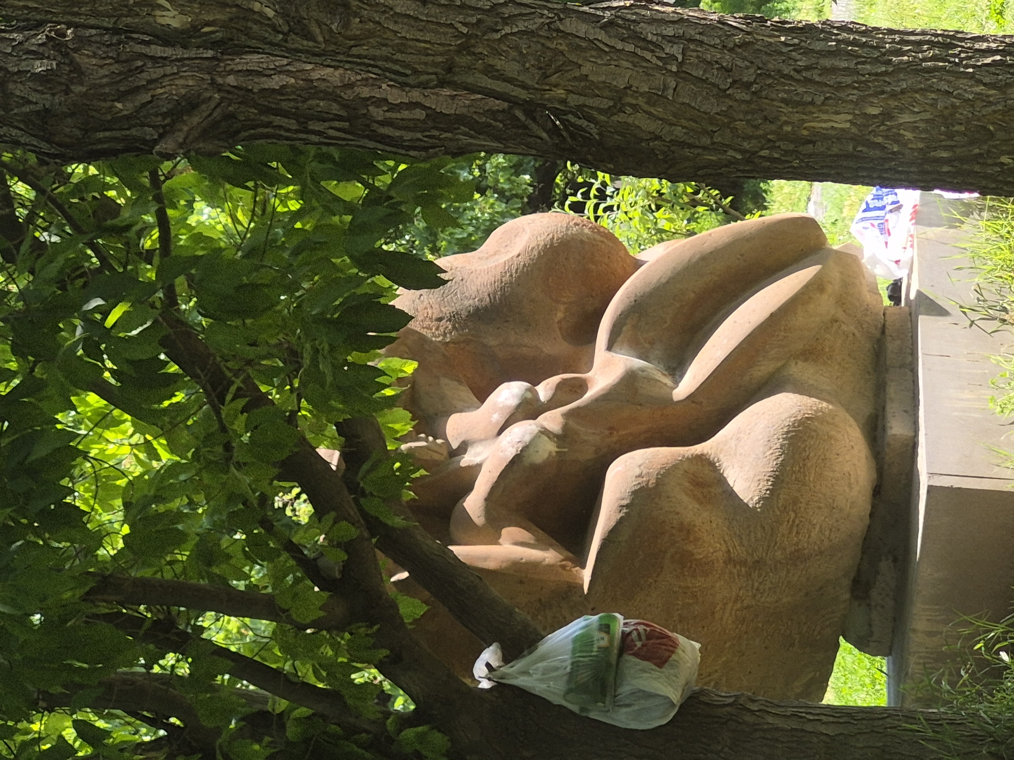 The image depicts an abstract sculpture in a park setting. The sculpture, made of a smooth, light-brown material, is a rounded, organic form suggestive of human figures. It is situated on a concrete pedestal. To the left of the sculpture, a plastic bag filled with items and tied with a string is hanging from a tree branch. Two trees frame the sculpture, one on the left with the bag hanging from it and another larger tree trunk on the right. The area around the sculpture is covered with grass and foliage, indicating an outdoor environment. In the background, more greenery and trees are visible. The lighting suggests it is daytime with sunlight filtering through the leaves of the trees. There are no people visible in the image. The scene appears to be in a public park, possibly in Yerevan, Armenia, given the presence of trees and an outdoor sculpture.