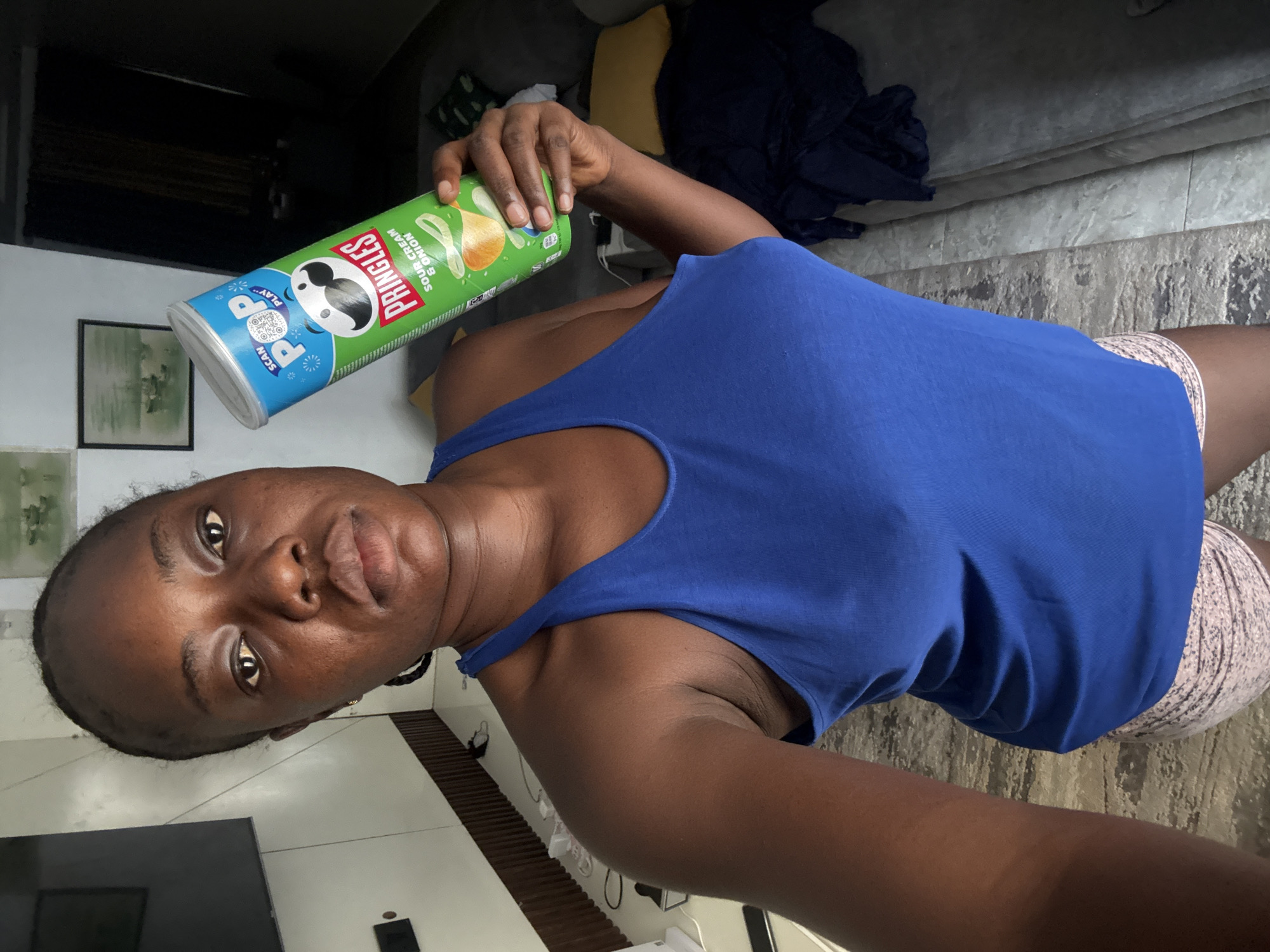 A dark-skinned woman in a blue tank top and patterned shorts holds a can of Pringles Sour Cream & Onion potato chips at arm's length. The location appears to be indoors, possibly a living room, with a television, framed picture on the wall, and a gray patterned rug visible in the background. There are objects of furniture that are positioned in the background, while the wall features a light gray color. The woman is the primary subject. There is no specific evidence in the image to suggest that the photo was taken in the city of Iddo Tudunwada, Nigeria.