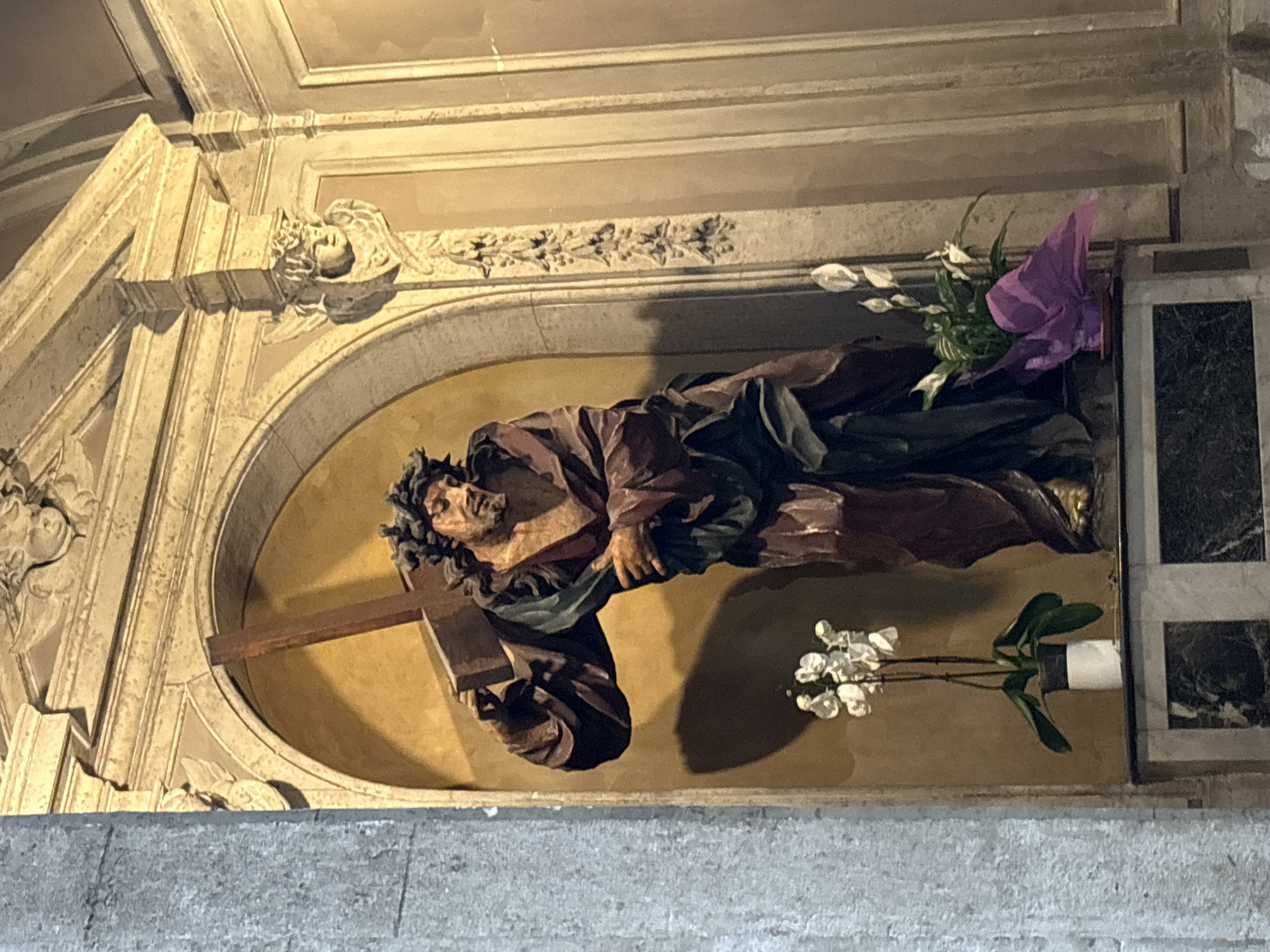 The image features a detailed statue of Jesus Christ, likely carved from wood, depicted in a moment of profound suffering as he carries the cross. He is shown with a pained expression, a crown of thorns on his head, and flowing robes rendered in dark red and green hues, struggling under the weight of the rough wooden cross.
The statue is housed within an arched niche, set against a warm, golden-brown background. This niche is part of an ornate architectural setting, characterized by intricately carved stone or plasterwork. Above the niche, there are decorative moldings, including a corbel featuring a carved angelic head with wings, and a frieze adorned with botanical motifs. The arch itself is also decorated with patterns.
Within the niche, beside the statue, are two arrangements of flowers. To the upper right, a vibrant purple floral arrangement with green leaves adds a splash of color. Below it, a pot containing delicate white orchids is visible. The bottom of the frame shows a rough, grey stone surface, possibly a floor or ledge.
The setting suggests an interior of a church or religious building, illuminated by artificial light that creates shadows, enhancing the depth and drama of the sculpture. The artwork conveys a solemn and devotional atmosphere, characteristic of religious art found in places like Rome, Italy. There is no visible text in the image.