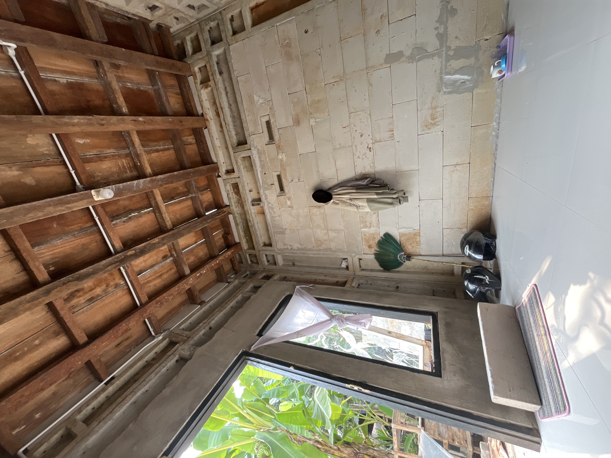 The image depicts an interior view of an unfinished building, possibly a house or storage area, with an open doorway revealing lush green foliage and banana trees outside. The ceiling is constructed with exposed wooden beams and planks. The walls are made of light-colored concrete blocks, with some areas appearing to be under construction or unfinished, showing patches of cement.
On the floor, which is tiled in a light, reflective material, there are several objects. Two black motorcycle helmets are placed side-by-side near the entrance. To the left, a long, rectangular concrete slab rests on a striped mat. A green fan made of woven material leans against the wall. A beige garment, possibly a jacket or hoodie, is hanging on a hook on the wall. Further to the right, on the floor, is a small purple tray holding a white cup and a blue lid or small bowl. The overall impression is of a functional space in progress, with elements of everyday life present alongside signs of ongoing construction.
The location is identified as Siwatu, Indonesia, suggesting a tropical or subtropical environment due to the prominent banana trees. The lighting suggests it is daytime, with sunlight casting shadows on the floor.