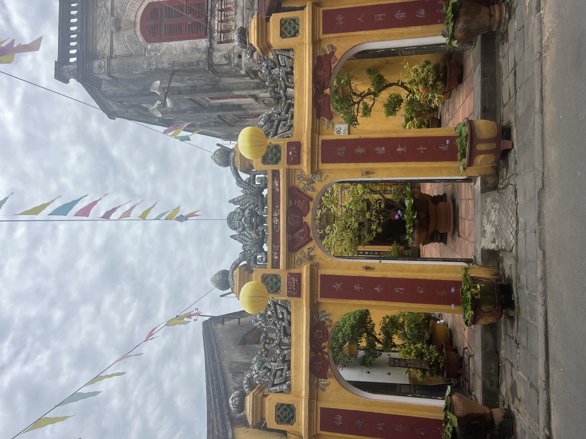 This image captures an ornate yellow gateway adorned with red calligraphy and intricate dragon-like sculptures, set against a cloudy sky. Colorful triangular flags are strung across the top, fluttering in the breeze. Large planters filled with lush green foliage and a few blooming lotus flowers flank the entrance. The gateway opens into a courtyard where more potted plants and trees are visible. The architecture suggests a traditional Vietnamese setting, likely in the ancient town of Hoi An, Vietnam, known for its historic buildings and vibrant culture. The overall scene conveys a sense of tranquility and cultural richness.