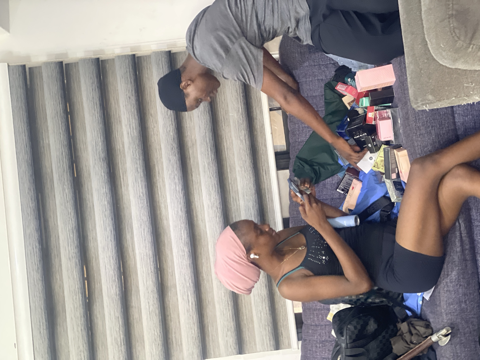 In a domestic indoor setting, possibly an apartment in Abuja, Nigeria, two individuals are seated on a purple couch. The individual on the left, a woman wearing a pink headscarf and a black tank top with a teal strap, is holding a smartphone and a light blue cylindrical object, possibly a cosmetic product. She is also wearing white earbuds and a necklace. The individual on the right, a man wearing a black beanie and a grey t-shirt, is reaching towards a pile of assorted items on the couch.
The couch is covered in various objects, including numerous small boxes in shades of pink, black, and white, suggesting cosmetic or perfume packaging, as well as a green bag and other unidentifiable items. To the far left of the couch, a black checkered bag and other personal belongings are visible. Behind the individuals, horizontal blinds cover a window, and a white wall is visible to the right. The overall scene suggests the unpacking or sorting of purchased items.