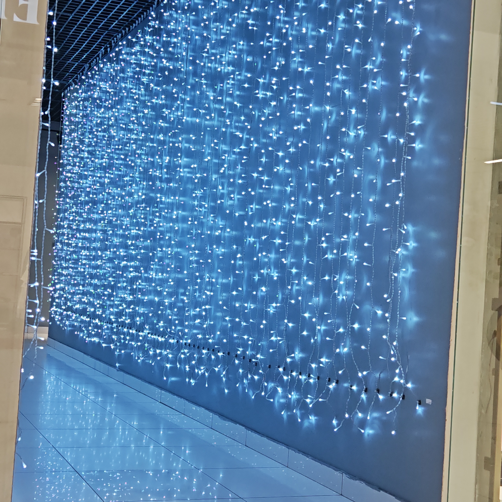 The image shows a wall adorned with numerous strings of blue LED lights. The lights appear to be hanging from a grid-like structure at the top, creating a waterfall effect. The bottom of the light strings are attached to the wall, a dull bluish-gray color. The location appears to be indoors, with tile flooring reflecting the blue light. The image is taken at an angle, making the lighting appear to stretch along a portion of the wall. The location is Taganrog City District, Russia.