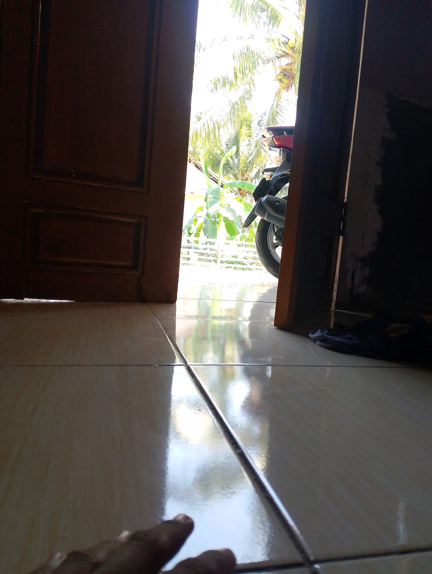 A low-angle shot captures the interior of a room looking out through an open doorway. The floor is covered in light-colored square tiles that reflect the sunlight streaming in. To the left, a dark wooden door is partially visible, with a panel design. The right side of the doorway is occupied by a dark, shadowed wall. Beyond the doorway, lush green foliage and palm trees with coconuts are visible, illuminated by bright sunlight. A portion of a motorcycle, primarily its rear wheel and exhaust, is parked just outside the doorway, partially obscured by the frame. In the foreground, a human hand is reaching into the frame from the bottom left corner, with its index finger extended. A dark piece of fabric, possibly clothing, lies on the floor near the right edge of the doorway. The scene appears to be a residential setting in Desa Babakan, Indonesia, given the architecture and tropical vegetation.