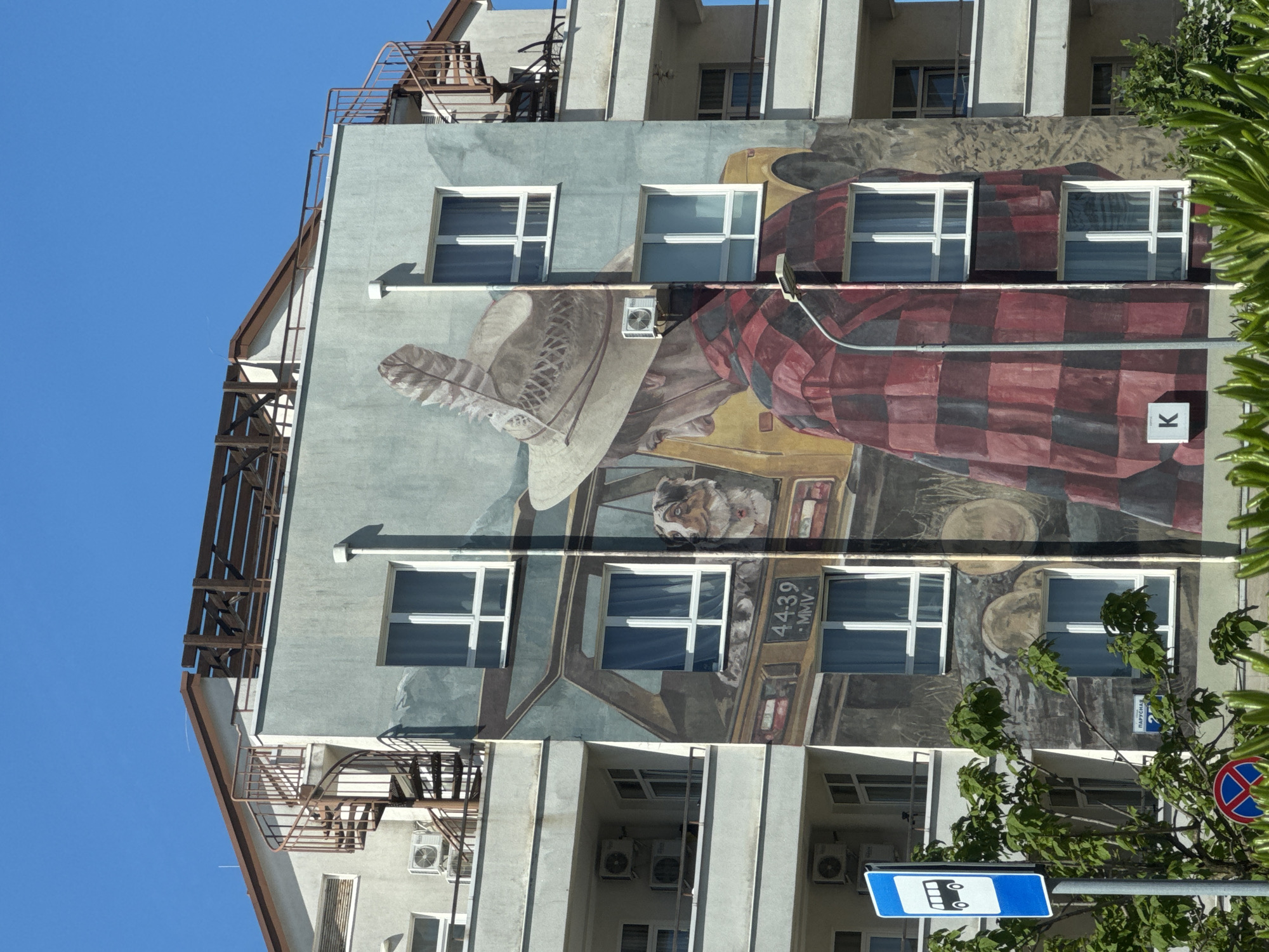 The original image is a vertical full shot of a building facade, rotated 90 degrees clockwise. When viewed upright, it depicts a multi-story building featuring a large, vibrant mural covering a significant portion of its side. The mural portrays a person wearing a wide-brimmed hat adorned with a feather, and a red and black checkered shirt or jacket. This figure appears to be in a vehicle, possibly a bus or truck, with a dog visible in the passenger seat area. Round, possibly barrel-like, objects are painted in the background of the vehicle. The building's actual windows are integrated into the mural, some appearing as windows of the painted vehicle.
The building itself is light grey or pale blue, with several white-framed windows evenly spaced across its stories. External metallic structures, possibly fire escapes or decorative elements, are visible on the left side of the building. Air conditioning units are mounted on the facade below the mural and on lower levels.
The scene is set under a clear, bright blue sky, indicating daytime and sunny weather. Green foliage from trees is visible in the foreground on the right side of the image (which would be the bottom left if rotated back to original orientation), partially obscuring the lower part of the building.
Visible text includes "4439 - MMV" painted on the mural near the dog's head, and a white square with a black "K" on the right side of the mural. Street signs are also present: a blue rectangular sign with a white bus icon (indicating a bus stop), a circular red and blue sign (likely a no-stopping/parking sign), and another sign with Cyrillic text "ПАРУСНАЯ 2" (PARUSNAYA 2). The location is Sirius, Russia.