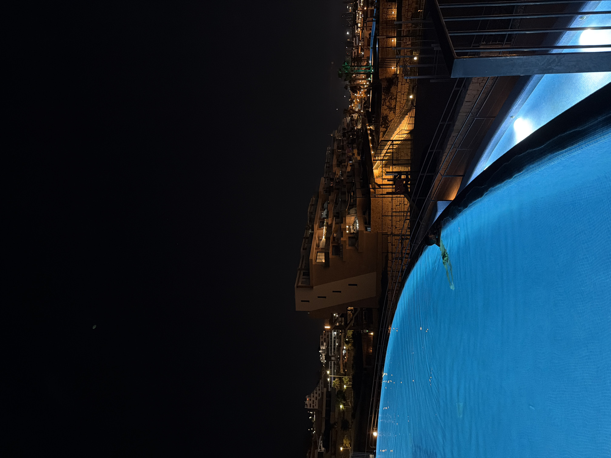 The image captures a nighttime view from an infinity pool overlooking the cityscape of Finestrat, Spain. The pool's vibrant blue water occupies the foreground, with its illuminated surface reflecting the ambient light. In the mid-ground, a multi-story building with angular architecture is visible, its facade illuminated by subtle lighting. Beyond this building, the city sprawls across the landscape, with numerous structures and streetlights creating a twinkling panorama. The sky is dark, indicating the late hour. A lone swimmer is visible in the pool, with their body partially submerged and their arms extended, suggesting they are either swimming or treading water. The overall scene conveys a serene and tranquil atmosphere, characteristic of a resort or residential area at night.
