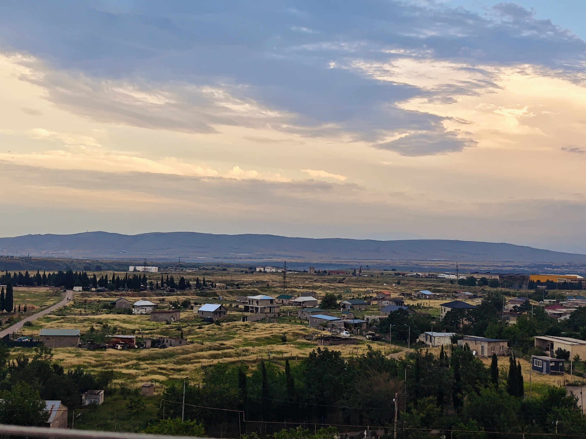 This is an eye-level, long shot, landscape photograph taken at dusk in Tbilisi, Georgia. The sky is filled with soft, pastel-colored clouds, with hues of blue, pink, and orange. Below the sky, a sprawling, semi-rural residential area is visible, characterized by numerous houses of varying sizes and architectural styles. The houses are interspersed with fields of dry, yellow grass, patches of green trees, and dirt roads. In the background, a hazy mountain range forms the horizon. The overall impression is one of a quiet, developing urban fringe at the end of the day.