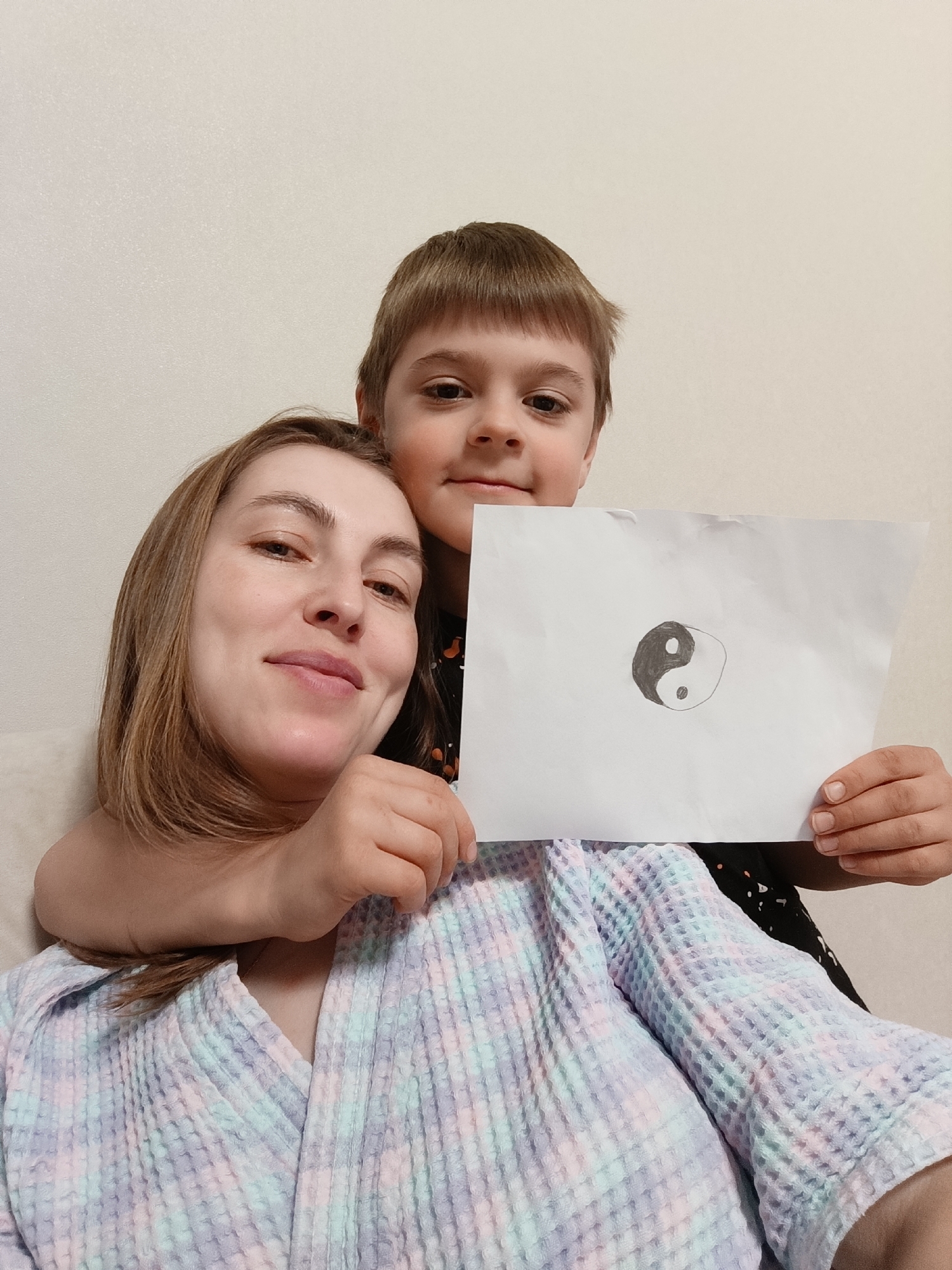 A mother and son are posing for a selfie. The son is behind the mother with his arm around her shoulder. The son is holding a drawing of a Yin and Yang symbol. The mother is wearing a pastel colored robe. They are indoors with a white wall in the background. The location is Chaikovsky, Russia.